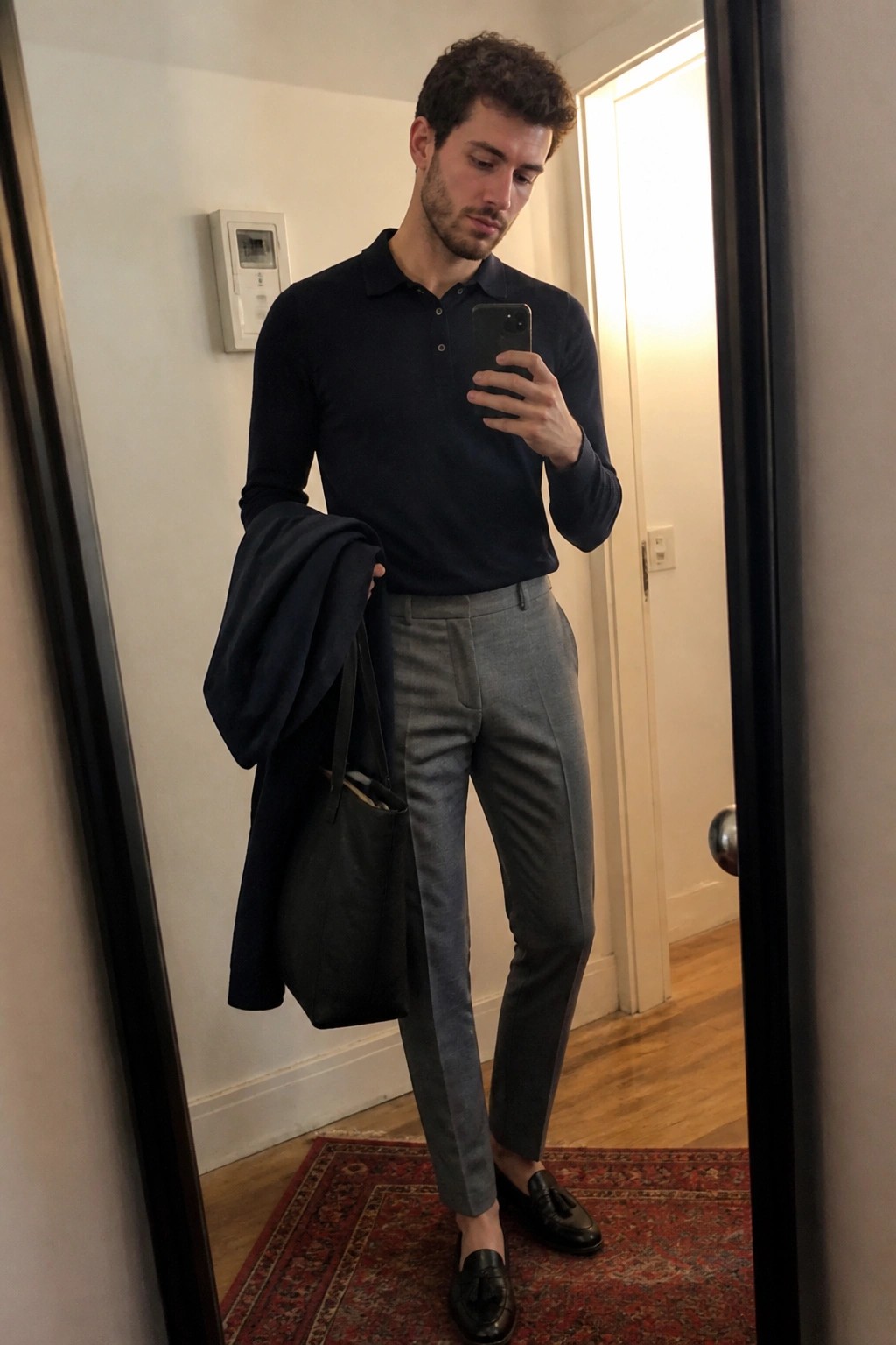 Man in navy long sleeve polo shirt tucked into gray wool trousers with black tassel loafers holding a navy coat and black tote bag while taking a mirror selfie