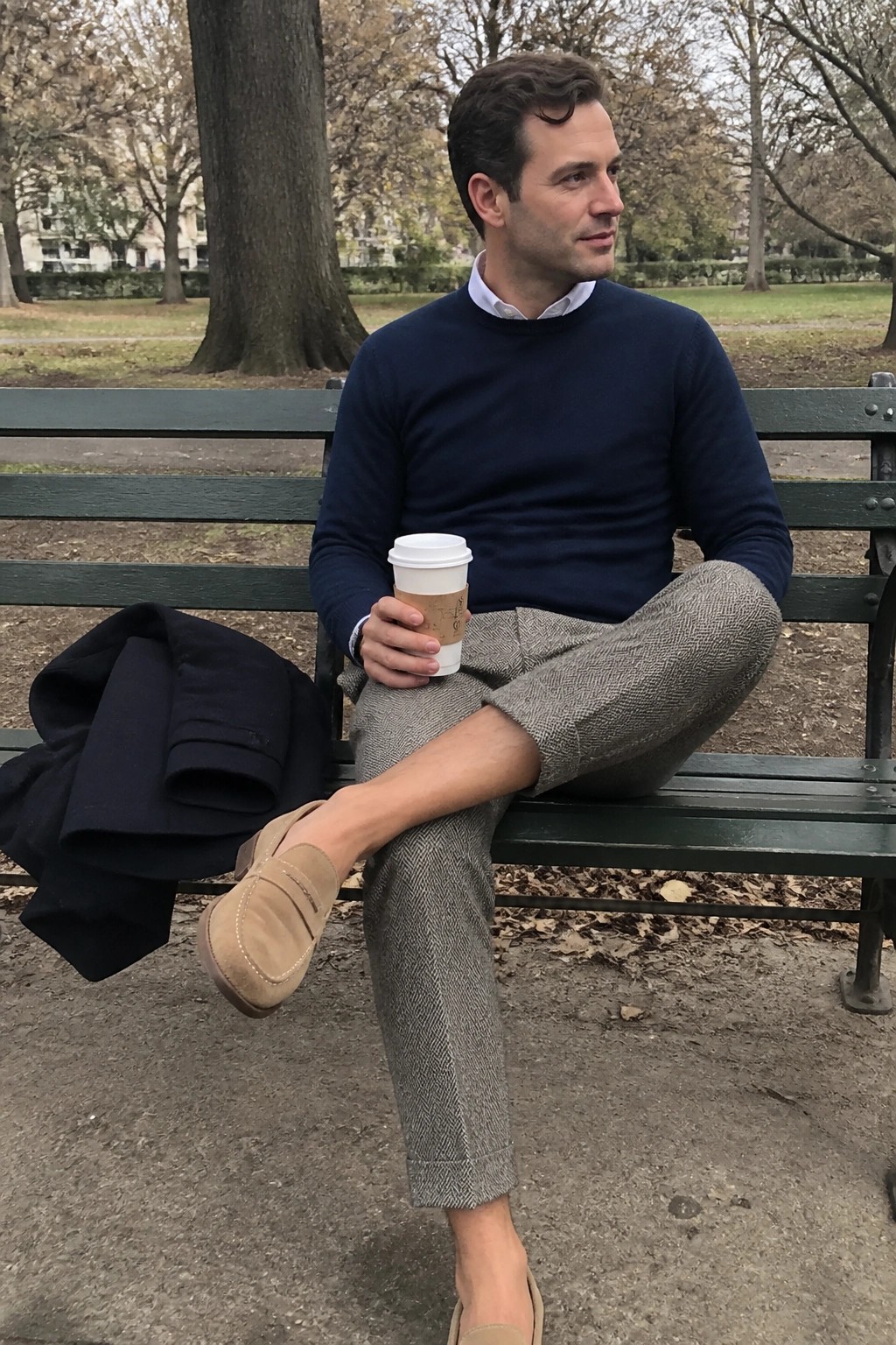 Man in navy sweater and herringbone pants sits on green bench holding coffee