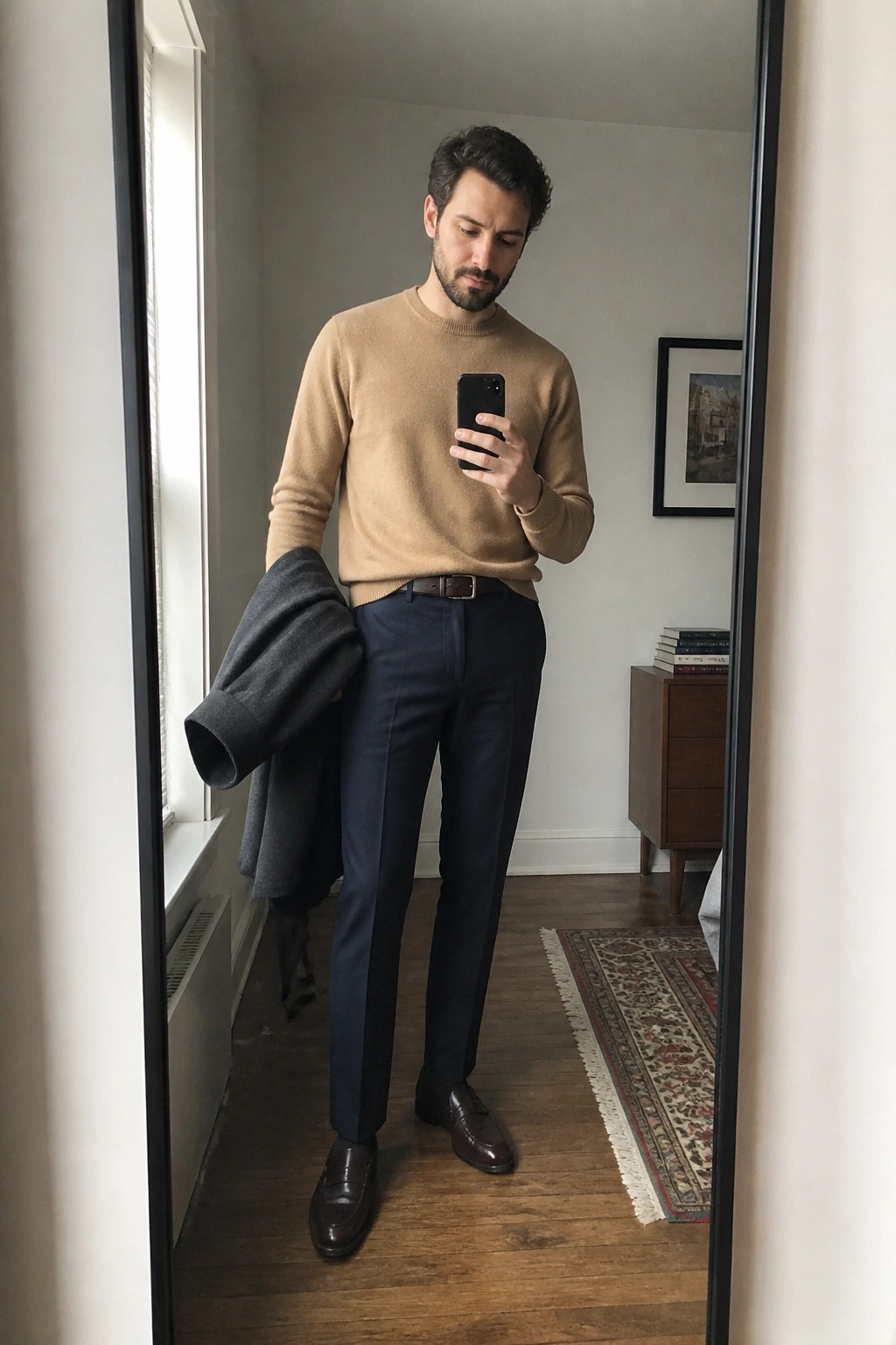 Bearded man in beige sweater and navy pants holds phone and gray coat in mirror.