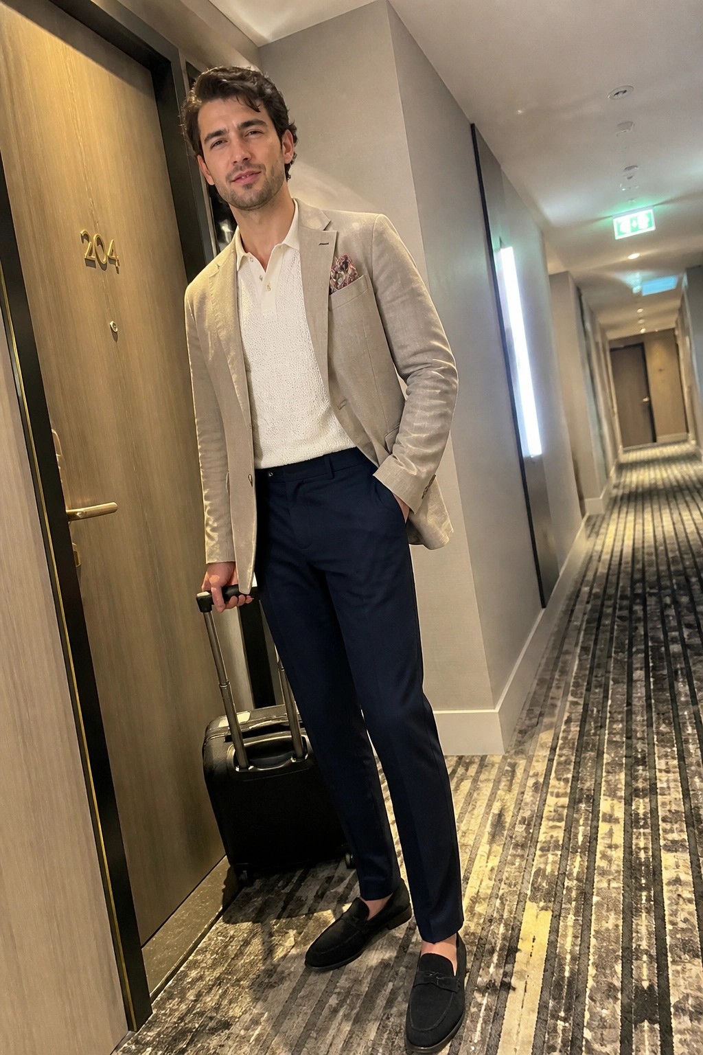 Man in beige blazer over white textured polo, navy wool trousers, black suede loafers and patterned pocket square standing in hotel hallway with black rolling suitcase.