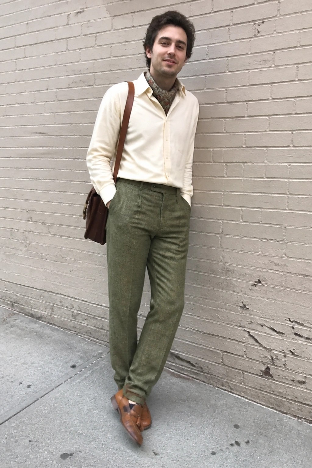 Man in cream shirt and green pants with brown bag by brick wall