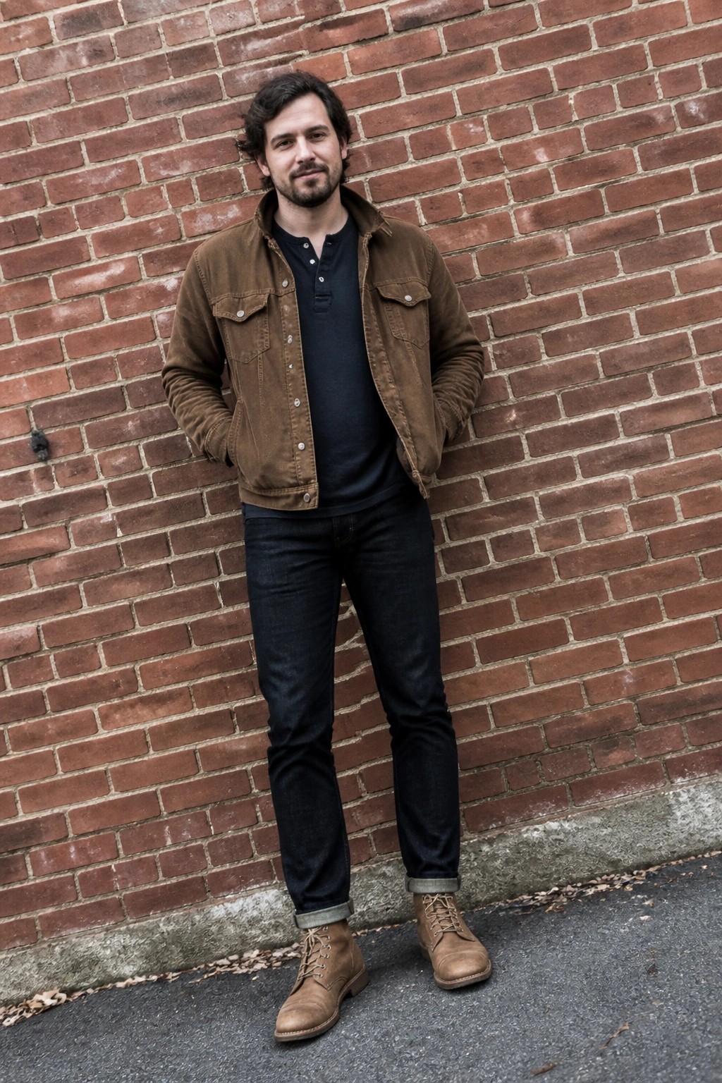 Man in brown jacket over navy shirt and dark jeans against red brick wall