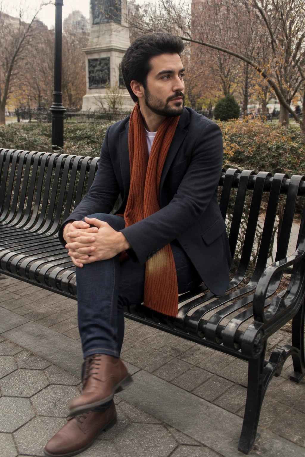 Man in navy blazer and orange scarf seated on black park bench, hands clasped.
