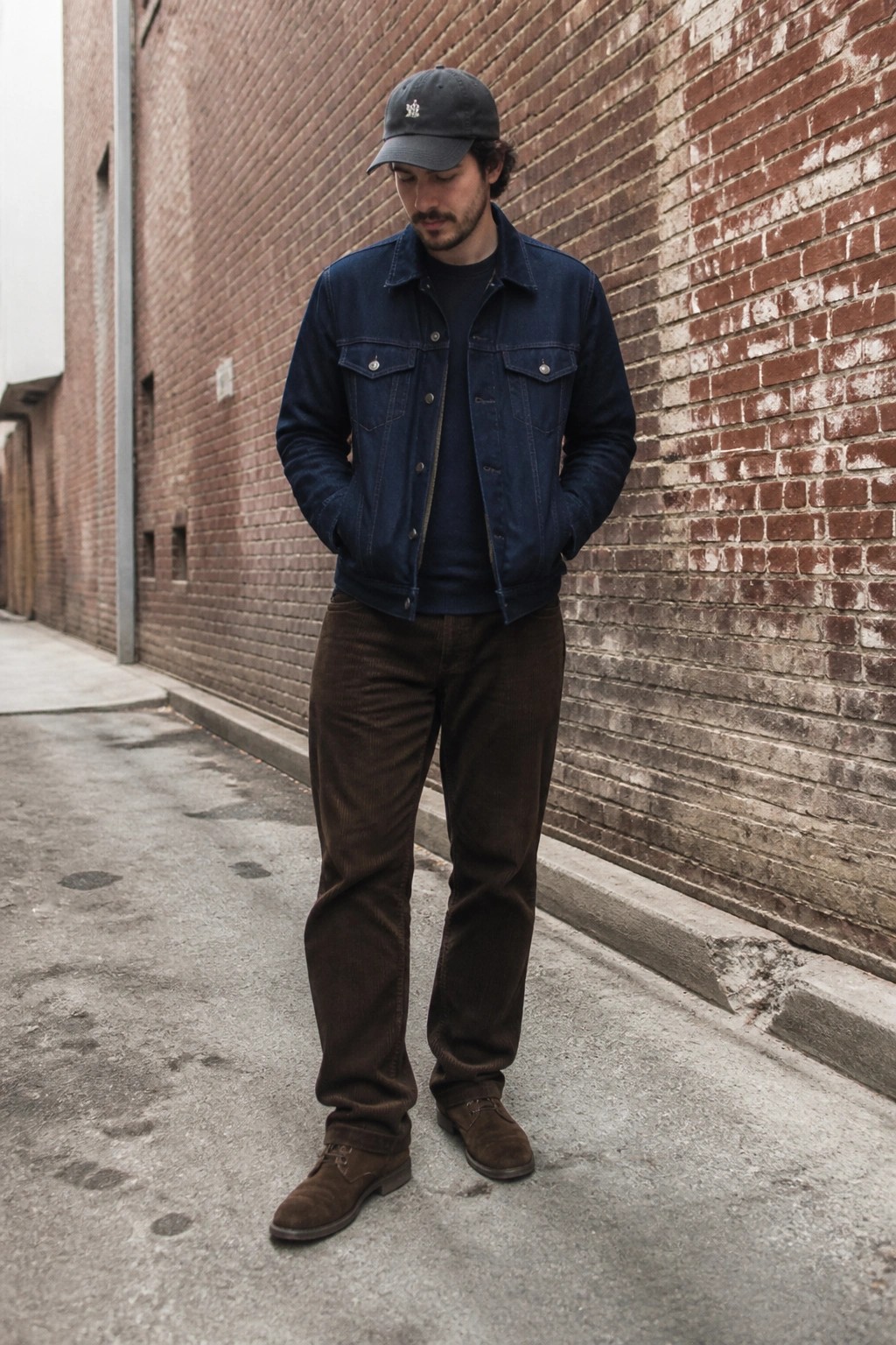 Man in blue denim jacket and brown corduroy pants stands in brick alley