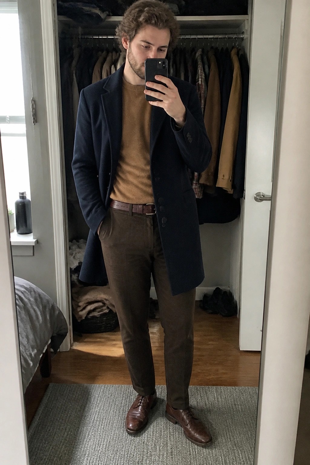 Man in navy overcoat and brown sweater taking mirror selfie in closet