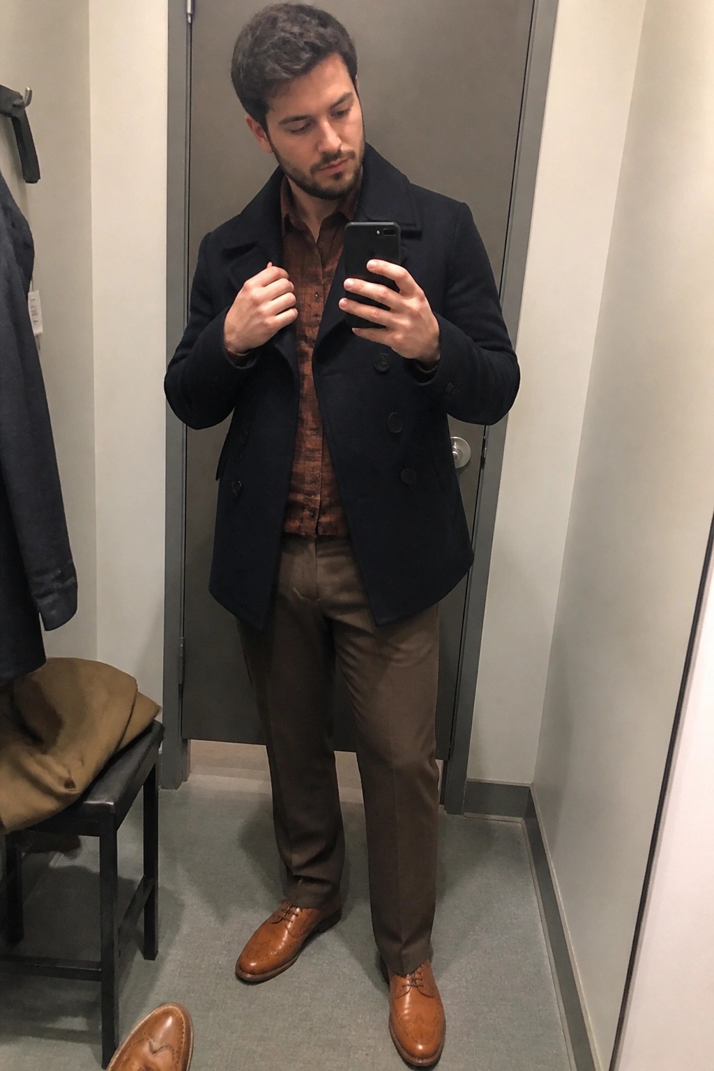 Man in navy peacoat over brown shirt and pants takes mirror selfie in fitting room