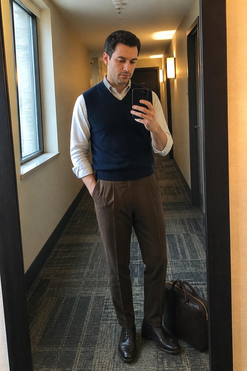 Man in navy vest and brown trousers taking mirror selfie in hallway with briefcase