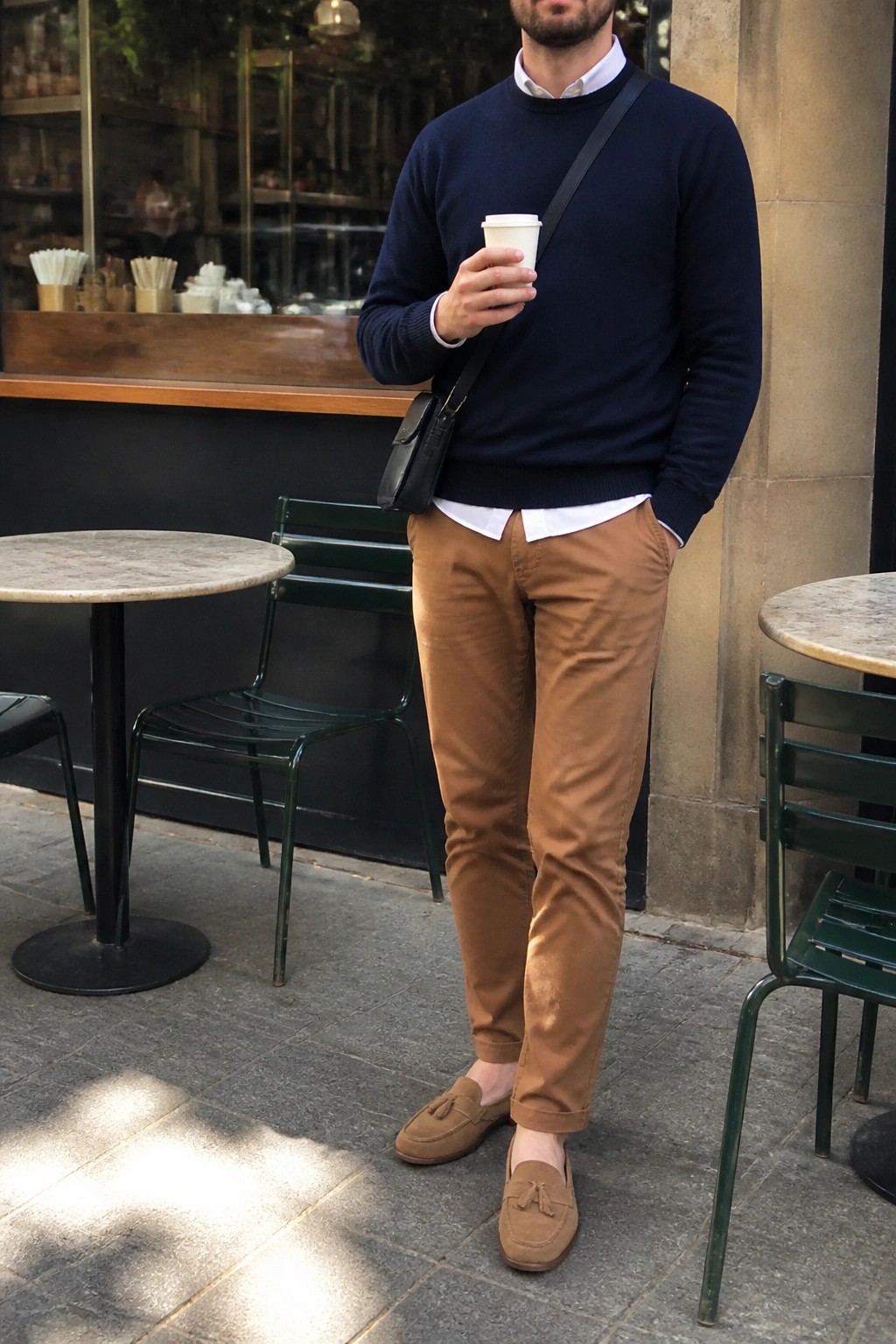 Man in navy sweater and brown pants holding coffee at outdoor cafe
