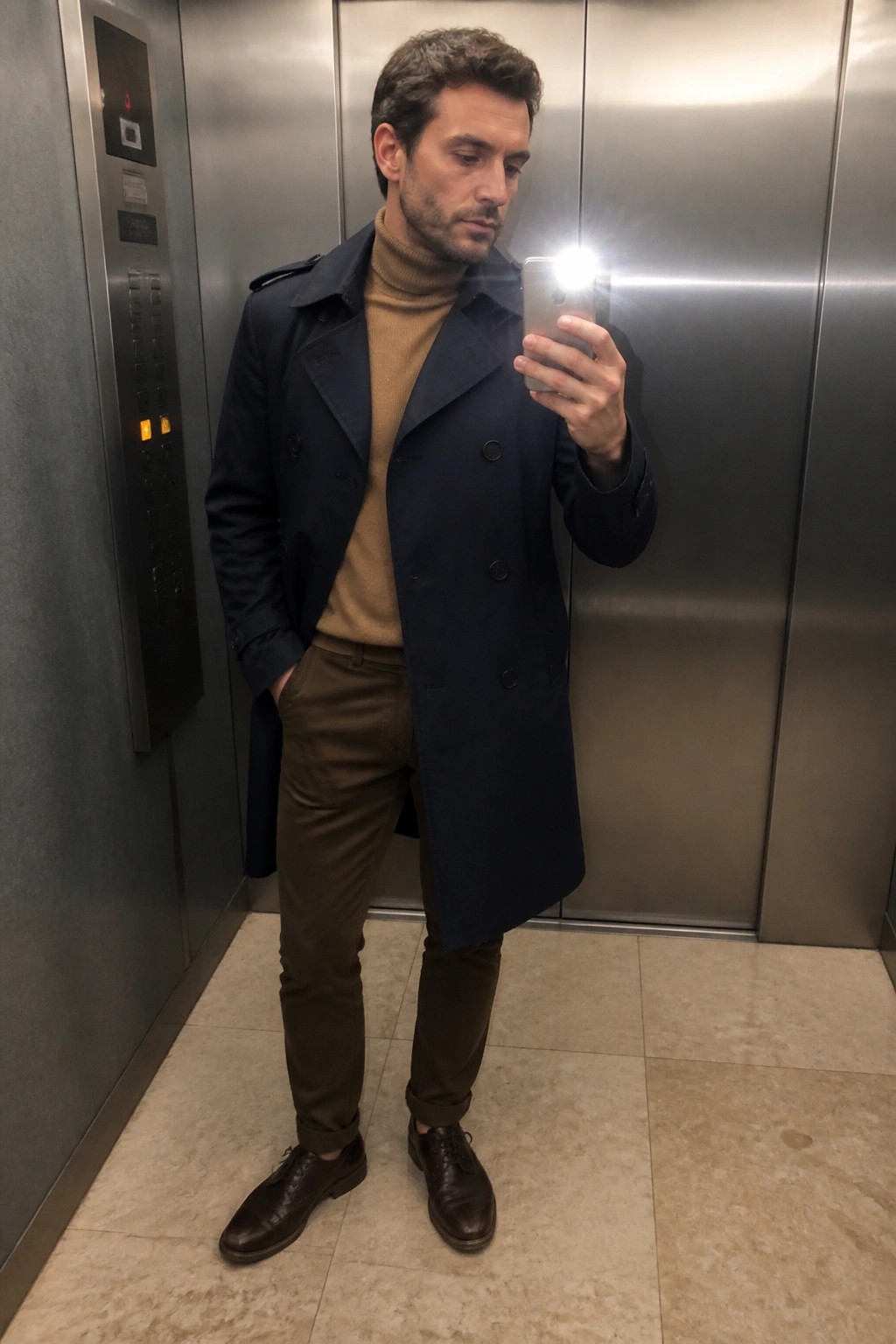 Man in navy trench coat and tan turtleneck taking selfie in elevator