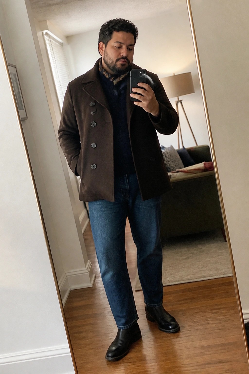 Man in brown coat over blue sweater and jeans taking mirror selfie with phone