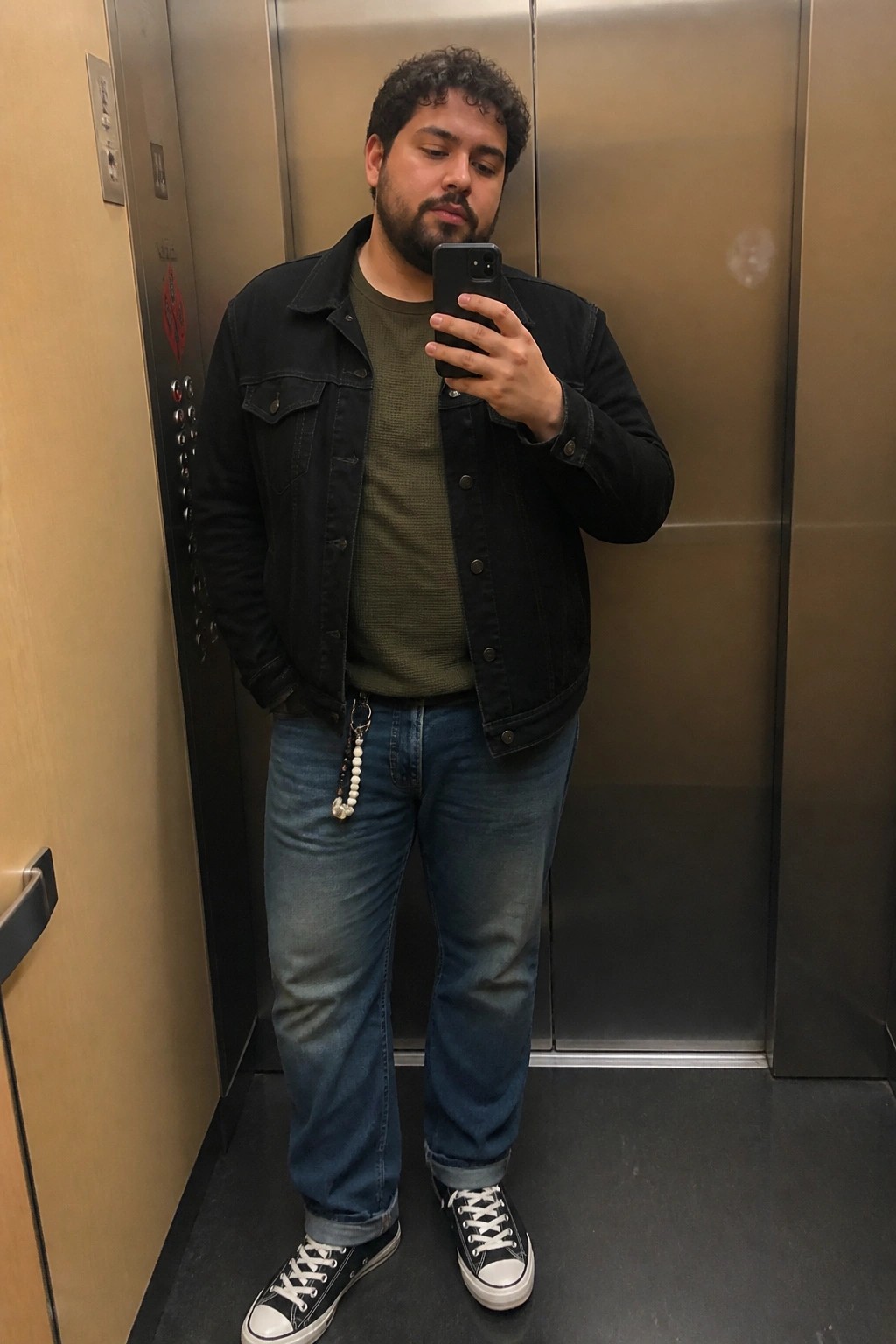 Plus size man in elevator mirror selfie wearing black denim jacket over olive green waffle shirt, relaxed straight leg blue jeans, black and white converse sneakers with beaded keychain on belt loop.