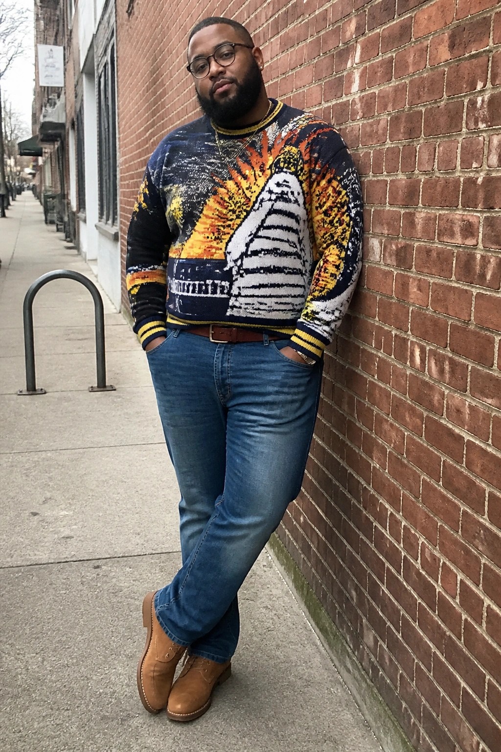 Bearded man in colorful graphic sweater leans against brick wall on sidewalk.