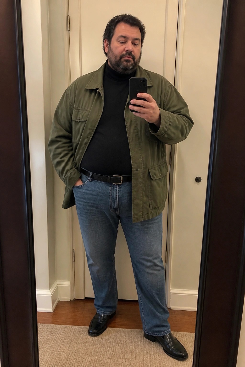 A plus size man wearing a green utility jacket over a black turtleneck, blue straight leg jeans with a black belt, and black loafers, taking a mirror selfie.