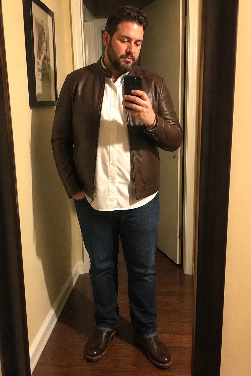 Man in brown leather bomber jacket over white button down shirt with blue straight leg jeans and brown brogue shoes taking a mirror selfie in a hallway.