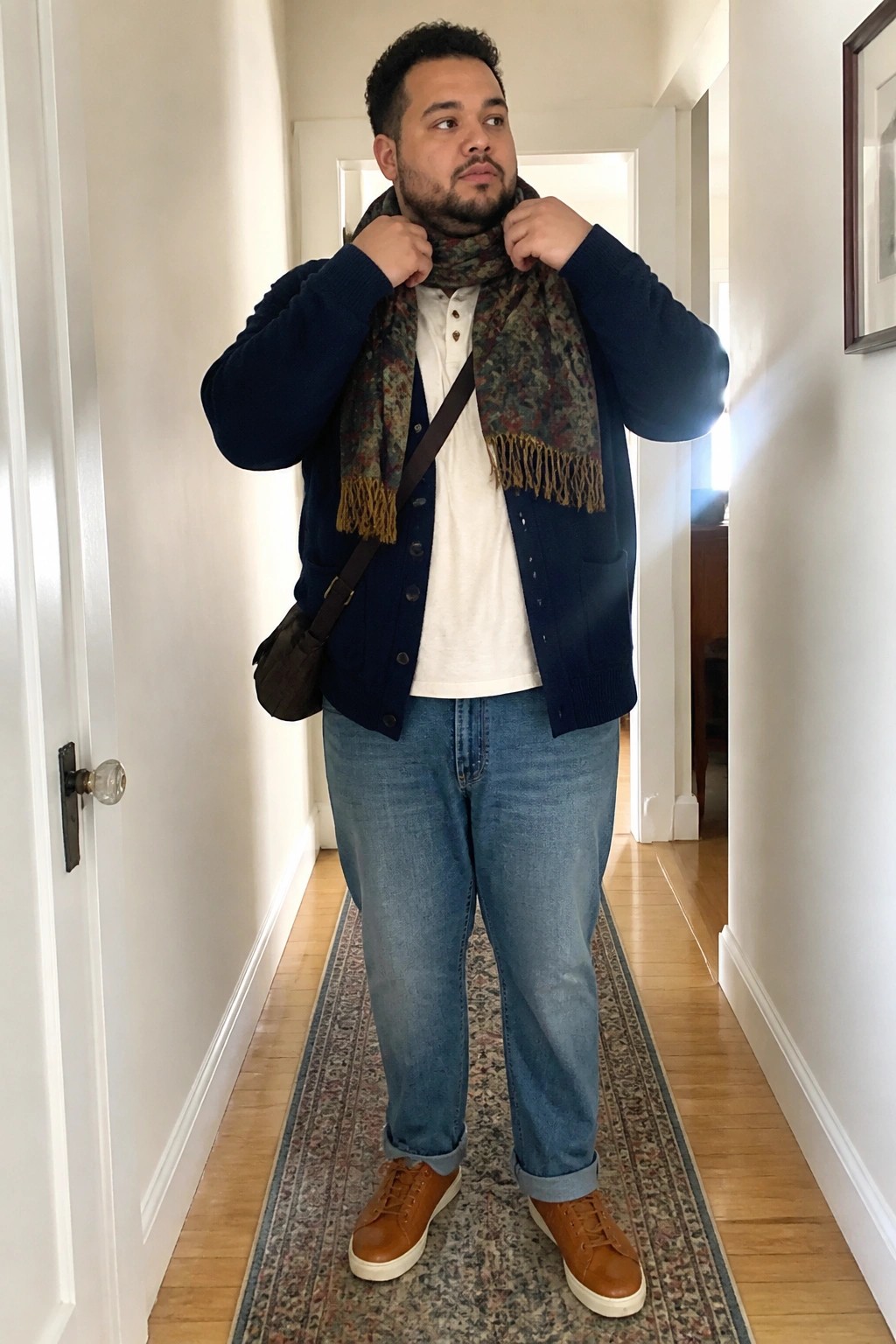Plus size man in navy cardigan over white henley shirt with patterned fringed scarf, relaxed straight leg blue jeans cuffed at ankle, brown sneakers and brown crossbody bag standing in hallway.