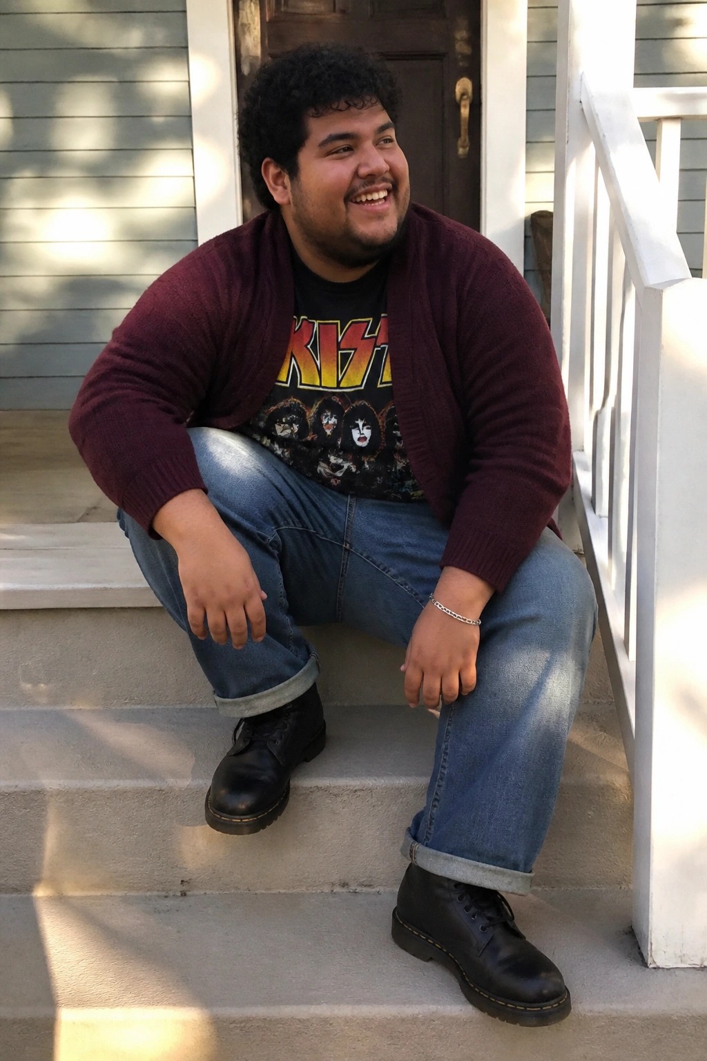 Plus size man wearing blue relaxed straight leg jeans cuffed at the ankle, black graphic KISS t shirt, maroon cardigan, black lace up boots, and silver chain bracelet while seated on outdoor steps.