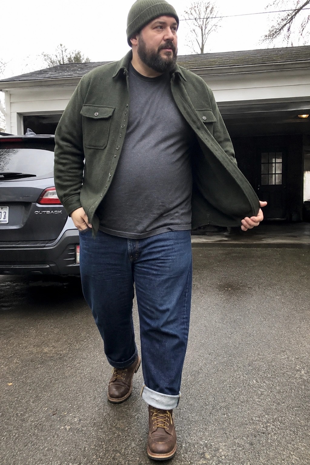 Plus size man wearing an open olive green wool shirt jacket over a gray t-shirt, paired with cuffed dark blue straight leg jeans and brown leather boots, plus a matching green beanie.