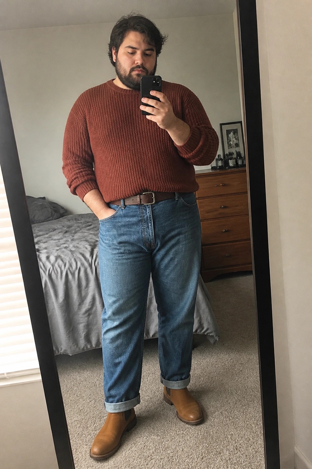 Bearded man in rust sweater and cuffed jeans taking mirror selfie.