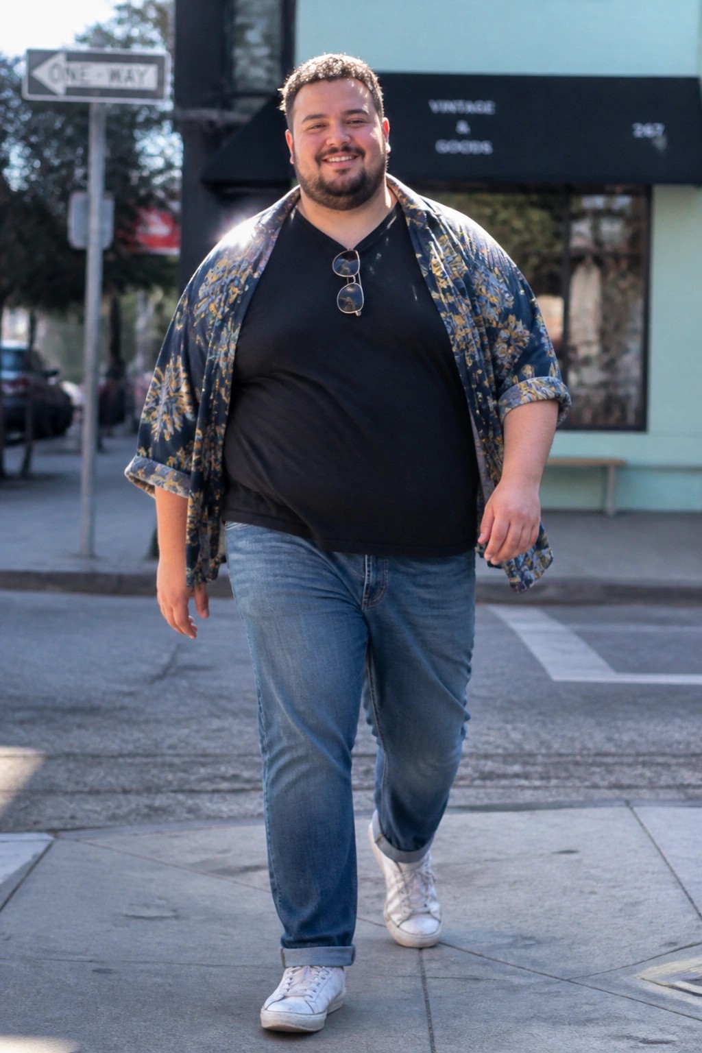 Smiling bearded man in floral shirt over black tee walks on city sidewalk