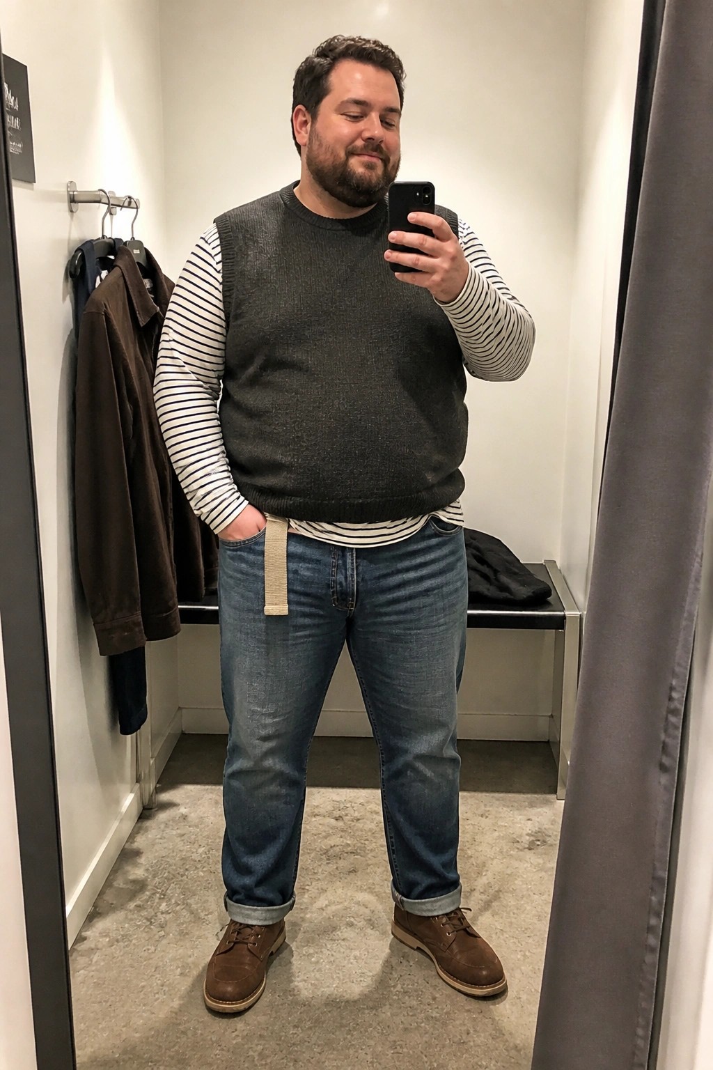 Plus size man in a gray knit sweater vest over a black and white striped long sleeve shirt tucked into relaxed straight leg blue jeans with a beige belt and brown lace up shoes.