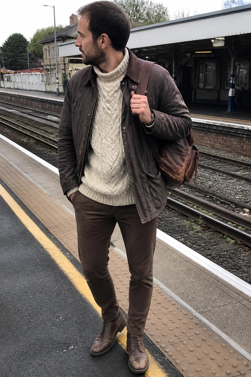 Man in brown leather jacket and cream sweater holding bag on train platform