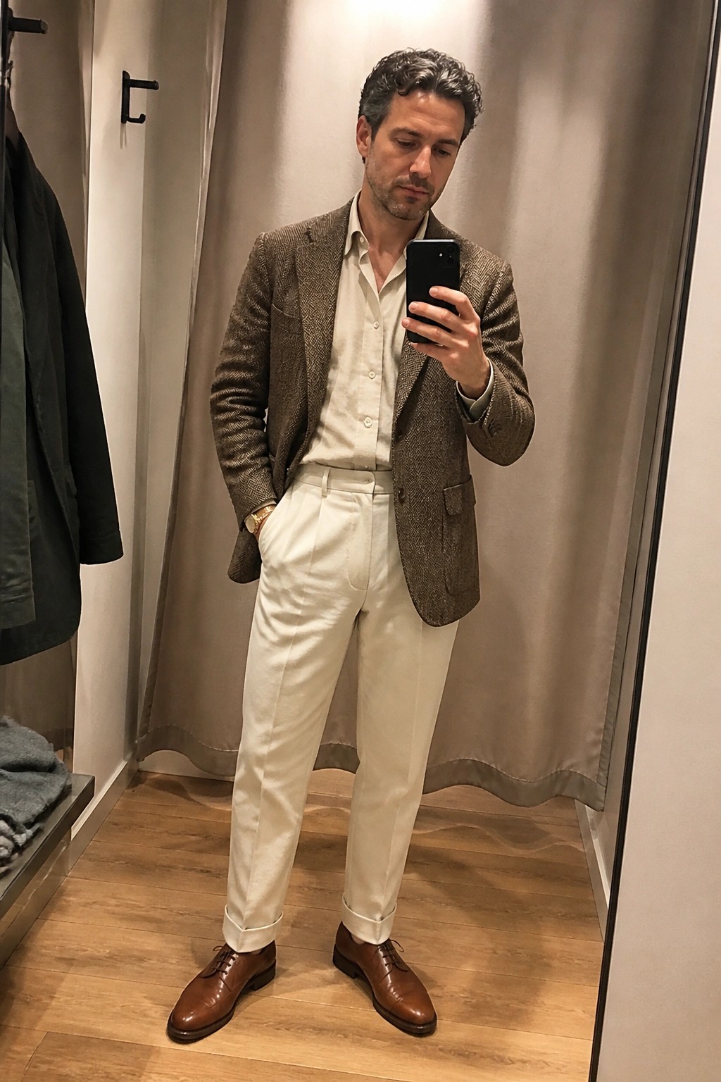 Man in a brown herringbone tweed blazer over a cream button down shirt paired with cream pleated trousers and brown leather dress shoes while holding a phone in a fitting room mirror.