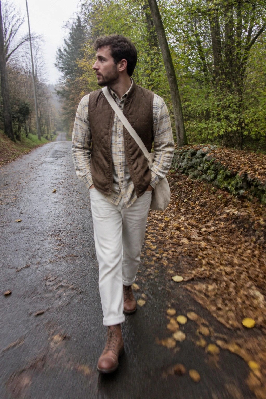 Man in plaid shirt and brown vest walks on wet autumn leaf road.