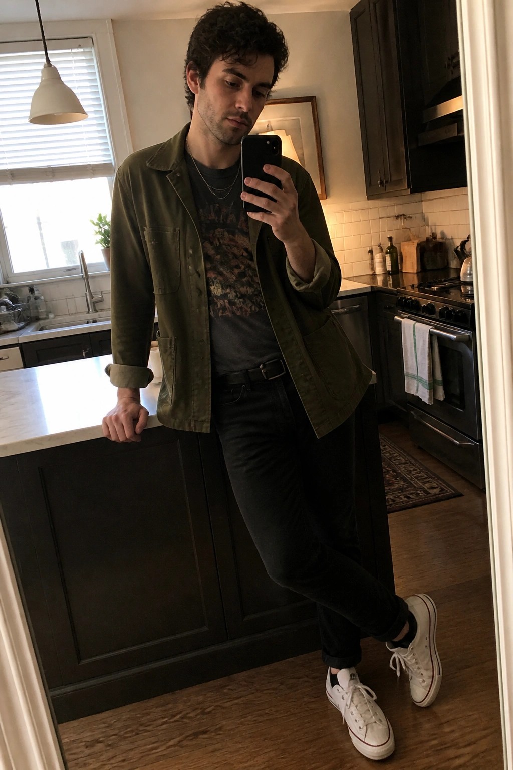 Curly-haired man in green jacket taking smartphone selfie in kitchen