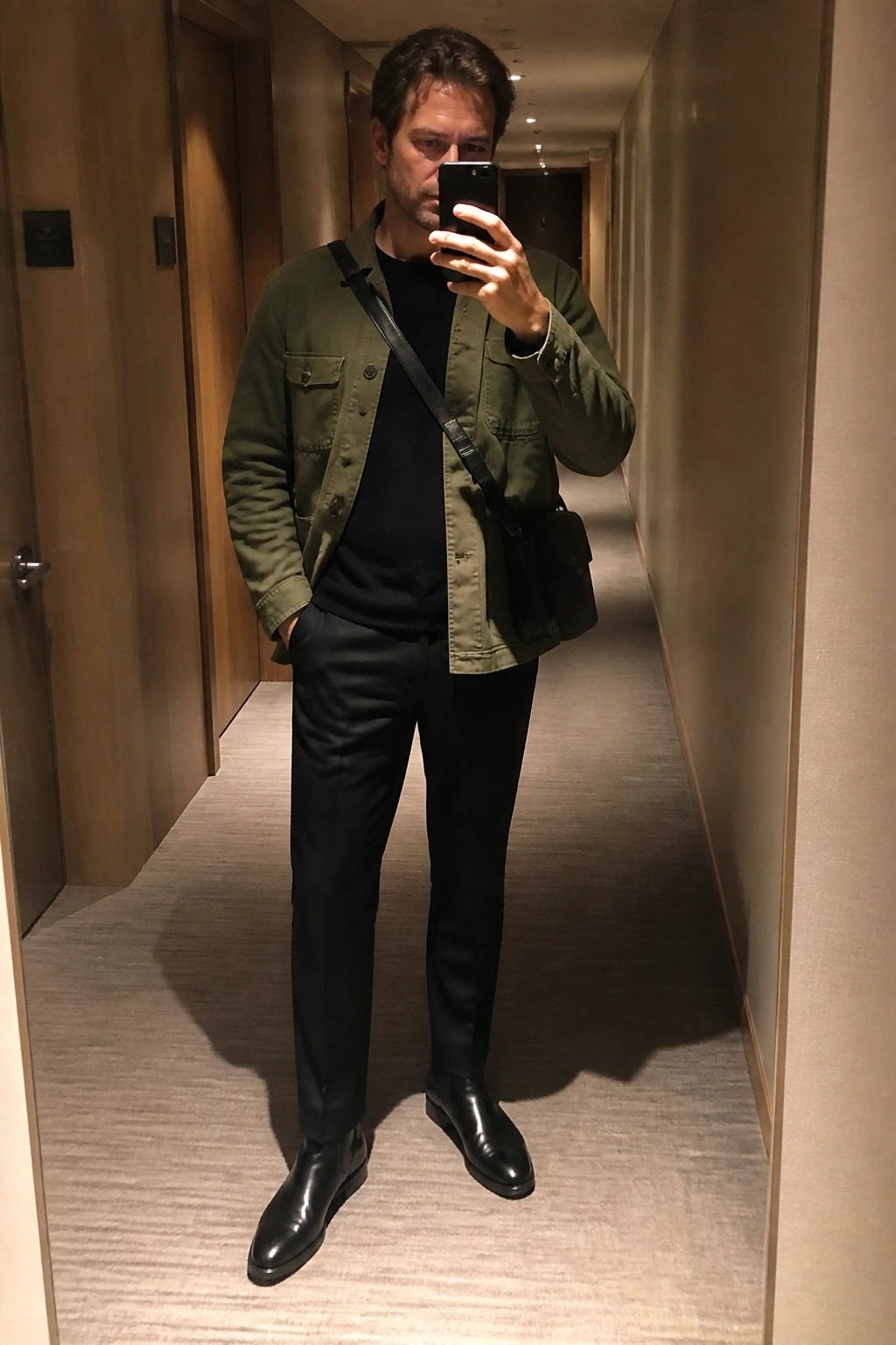 Man in olive green jacket taking mirror selfie in modern hallway