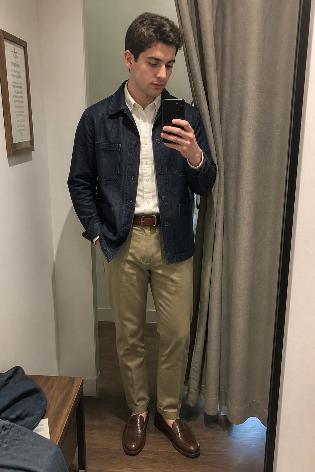 Man in dark blue denim chore coat over white button down shirt, khaki pants, brown leather belt and loafers, taking a mirror selfie.