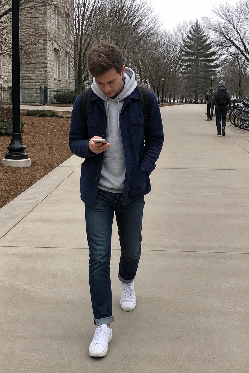 Young man in blue jacket over gray hoodie checks phone on campus walkway.