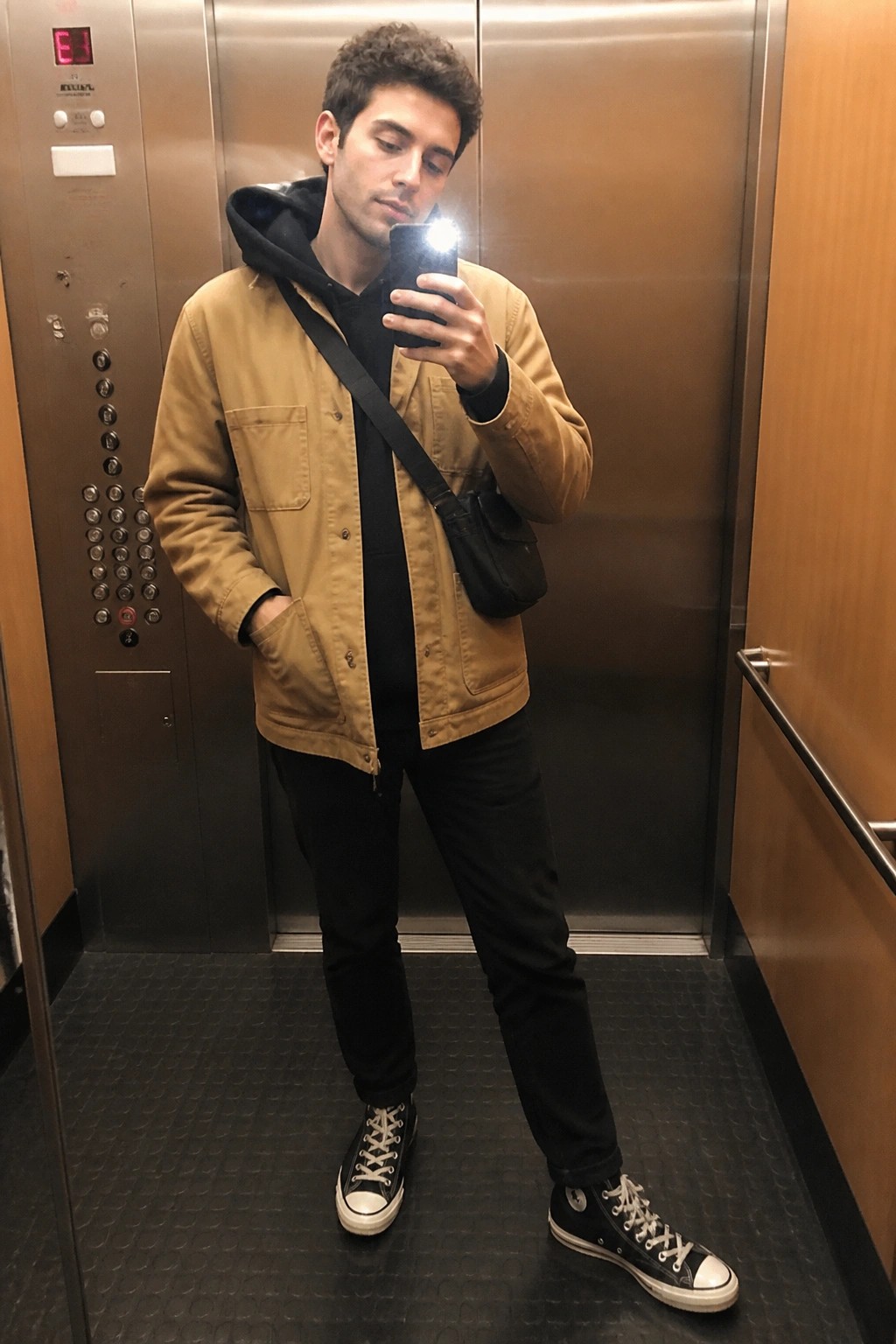 Man in tan jacket taking phone selfie inside elevator with black crossbody bag