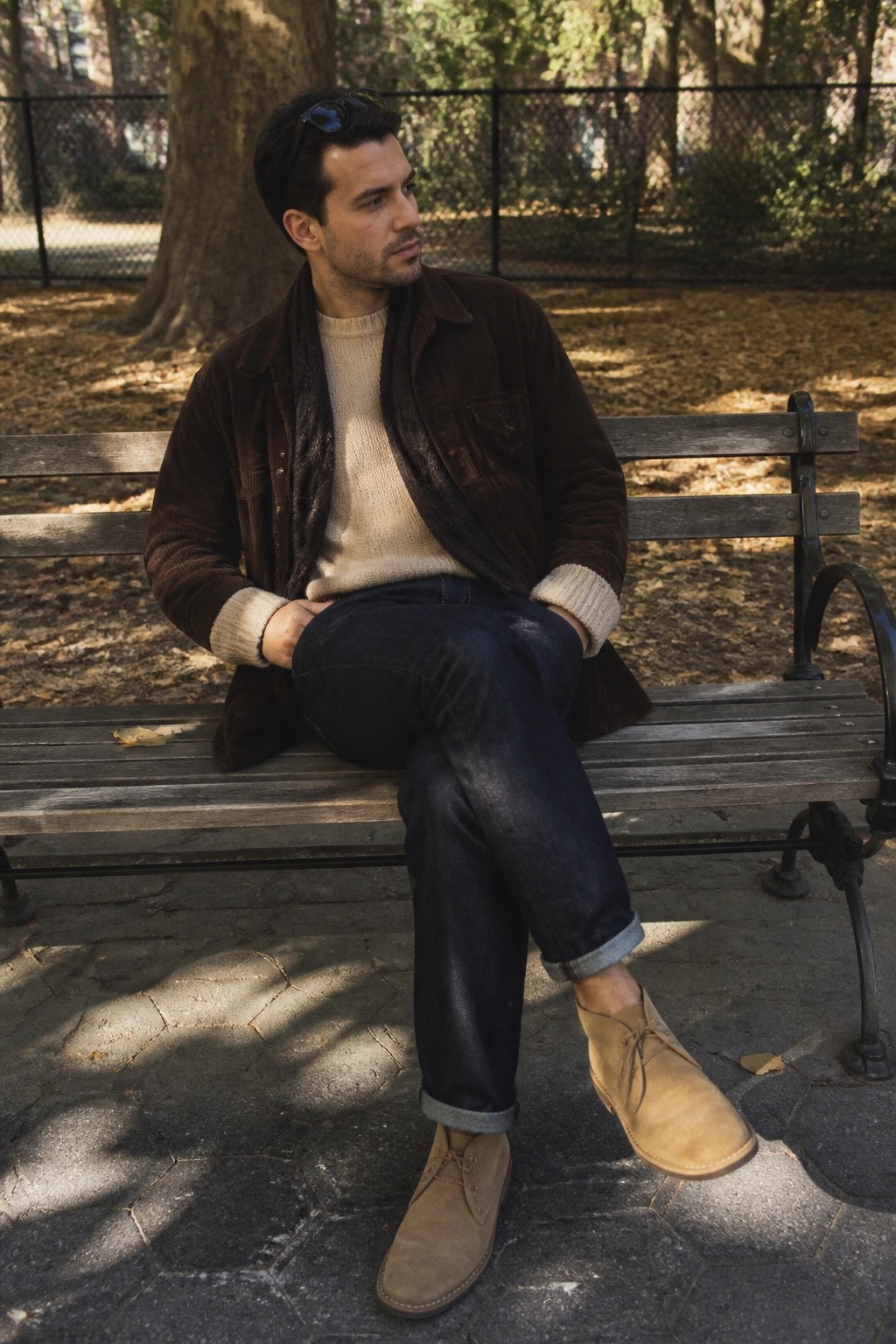 Man on wooden bench in brown corduroy chore coat open over cream knit sweater, dark cuffed jeans, and tan suede chukka boots with sunglasses resting on head.
