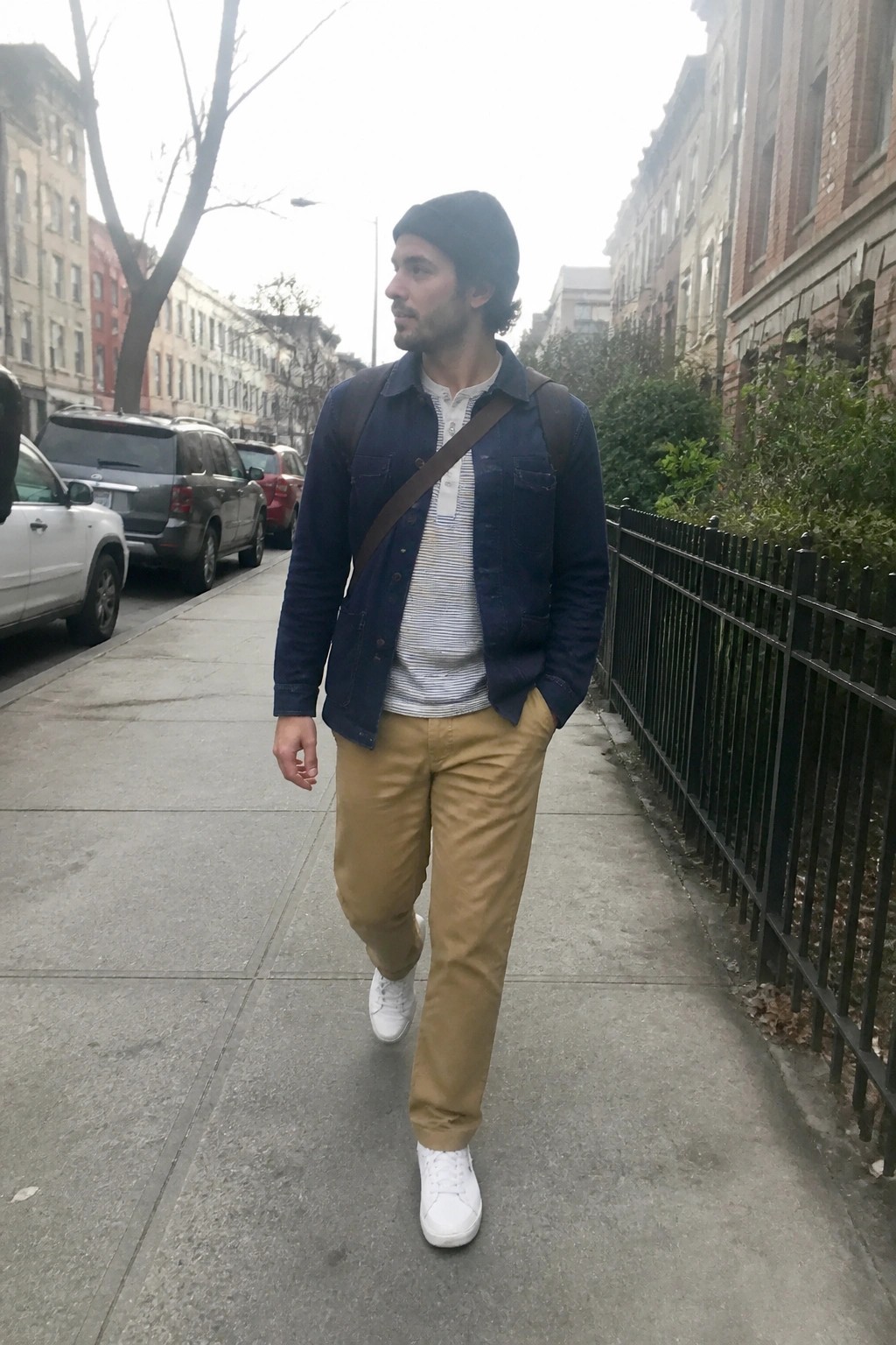 Man in blue jacket, beanie, and khaki pants walking city sidewalk with bag