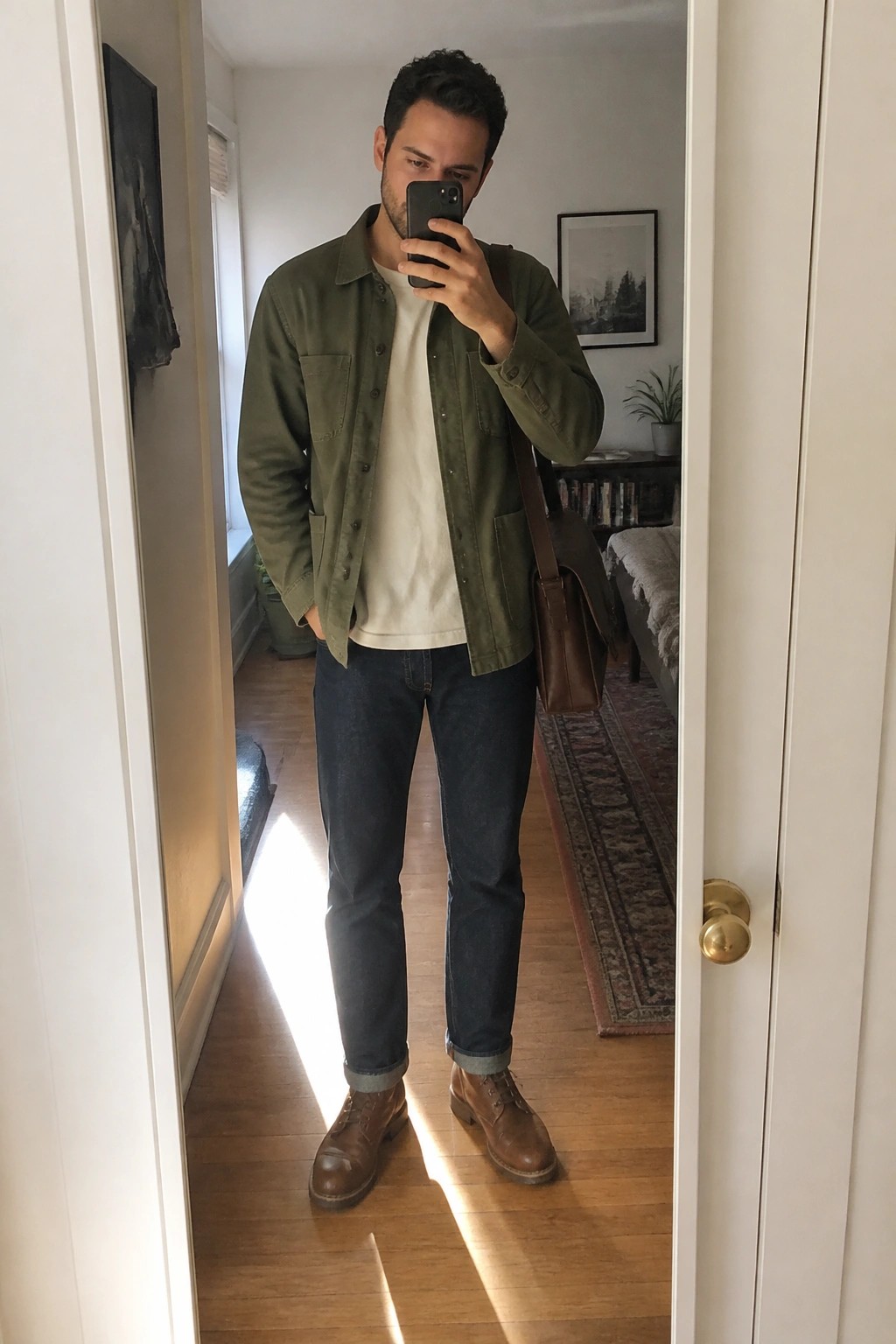 Man in green jacket over white tee with jeans and boots taking mirror selfie