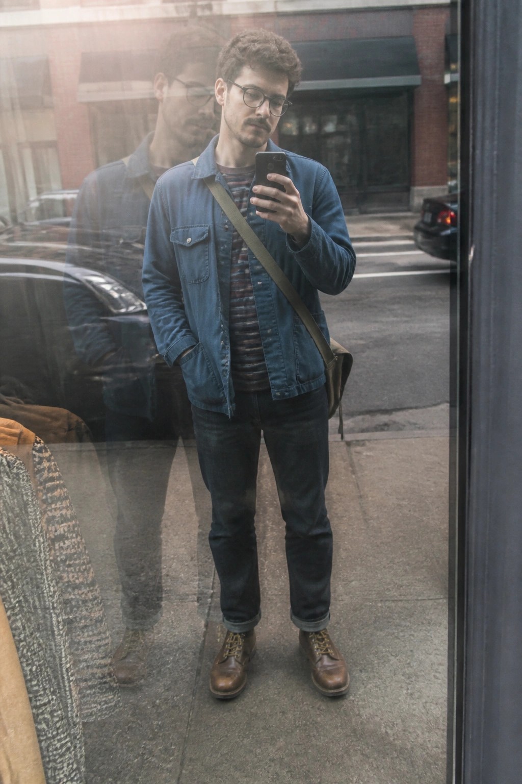 Man in blue denim jacket checks phone on city sidewalk with window reflection