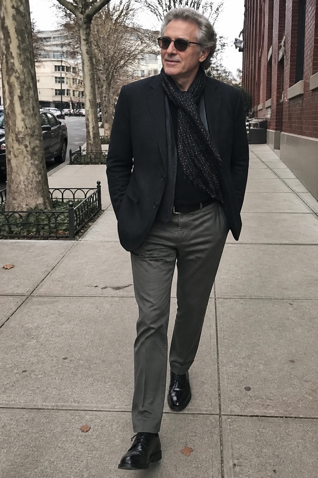 Gray-haired man in black blazer and patterned scarf walks on city sidewalk
