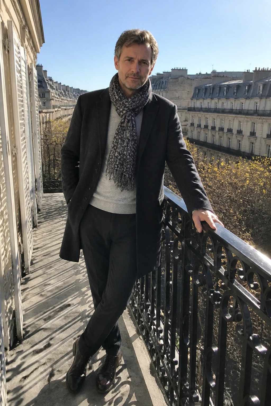 Man in black coat and patterned scarf on sunny balcony overlooking buildings