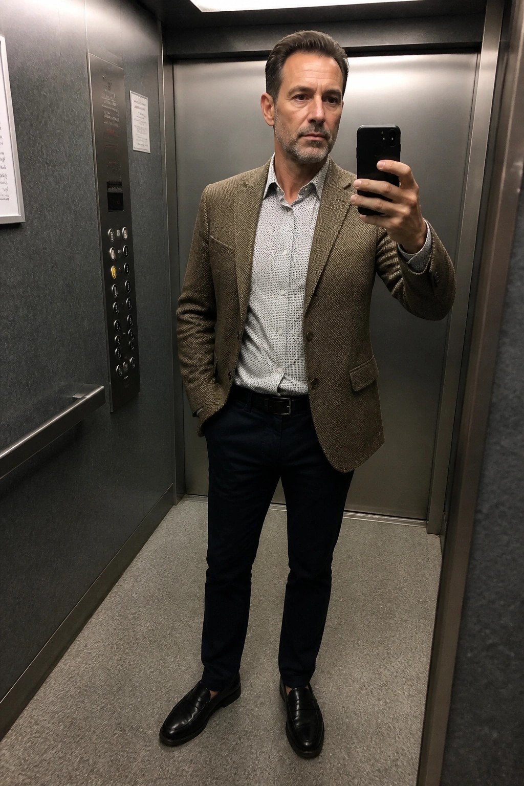 Man in brown tweed blazer takes selfie inside metal elevator with phone