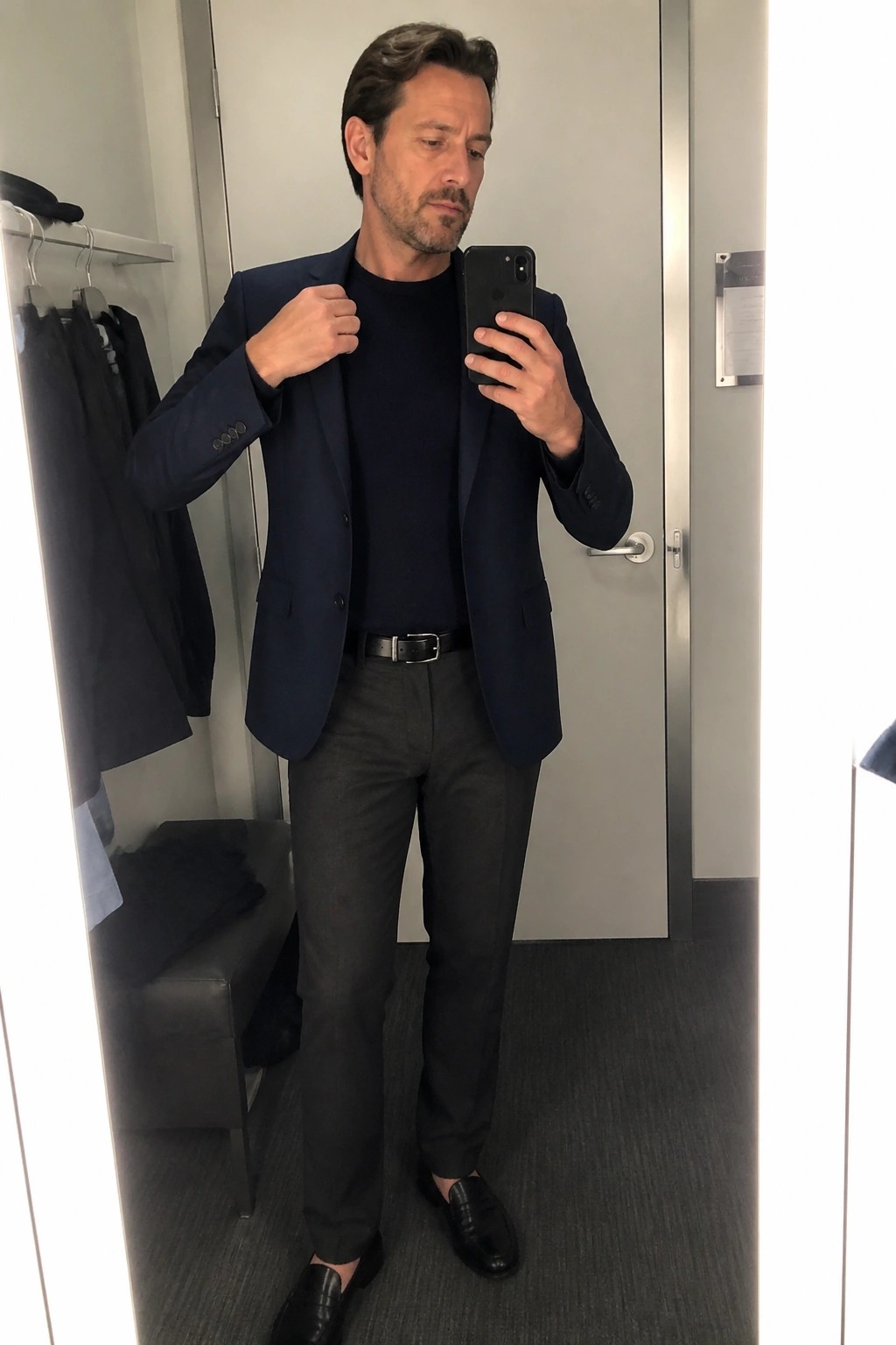 Man in navy blazer over sweater taking mirror selfie in fitting room