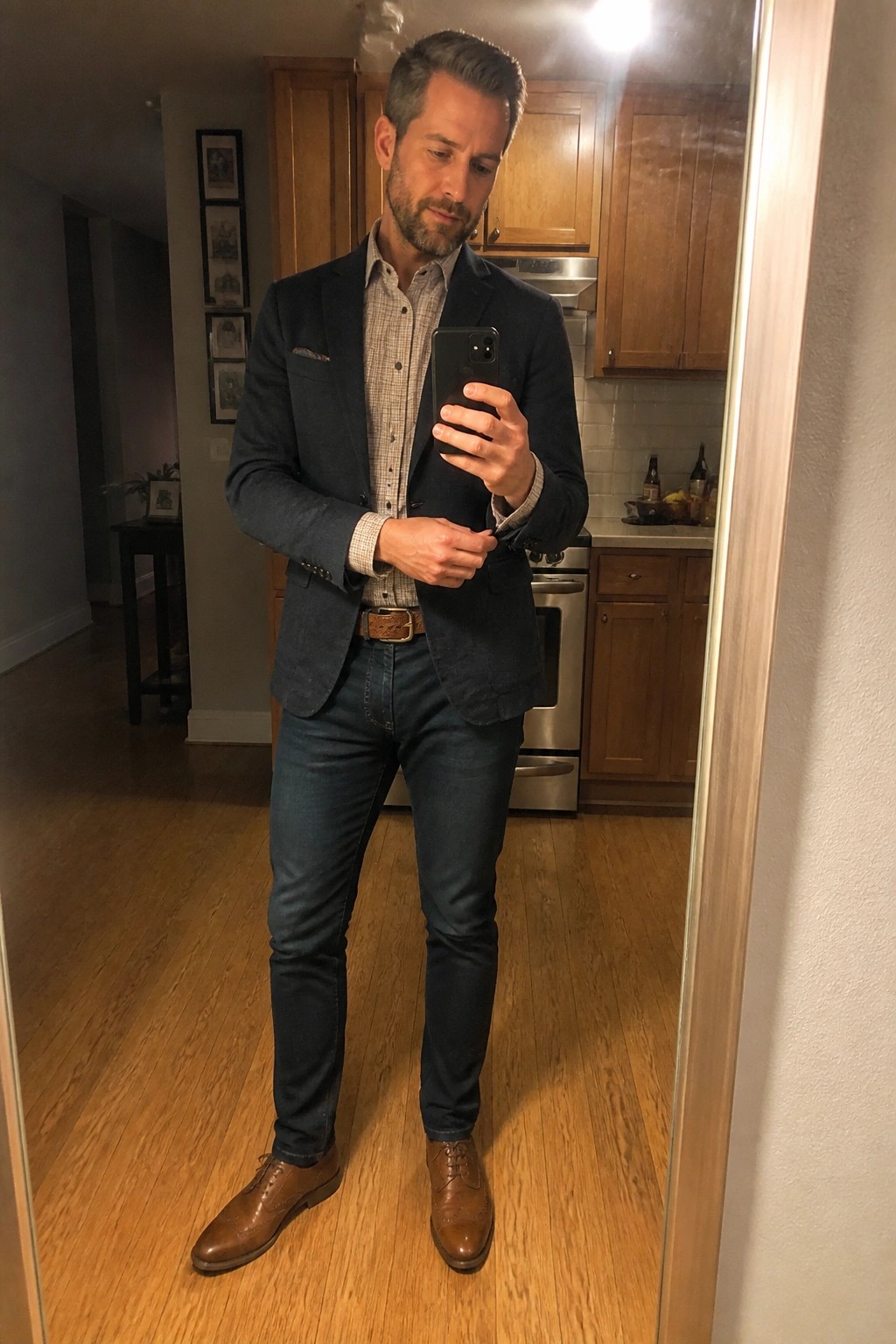 Man in navy blazer and jeans taking mirror selfie with phone in kitchen