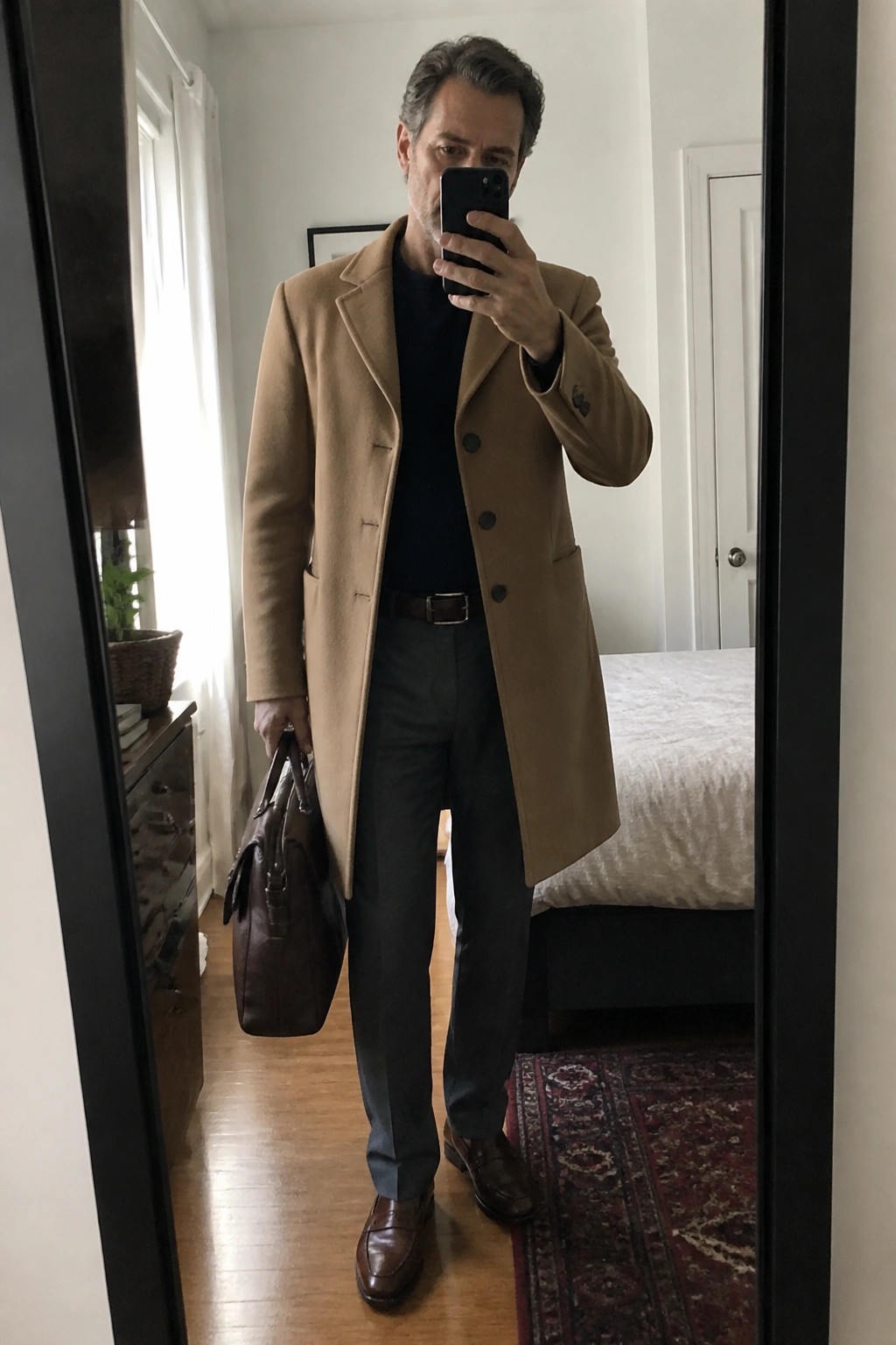Gray-haired man in tan overcoat and gray trousers holds leather bag for mirror selfie