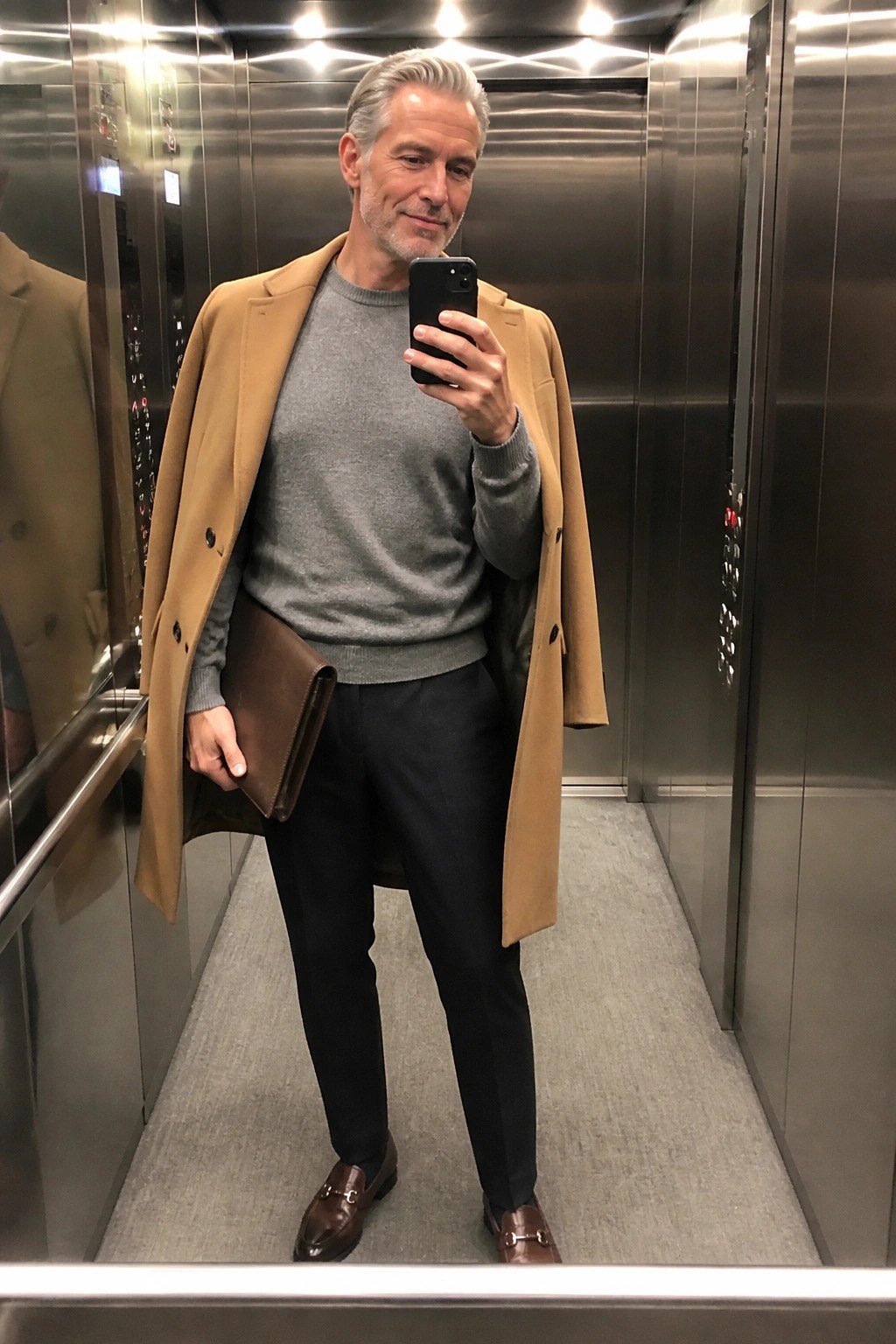 Man in tan overcoat and gray sweater taking elevator mirror selfie with phone