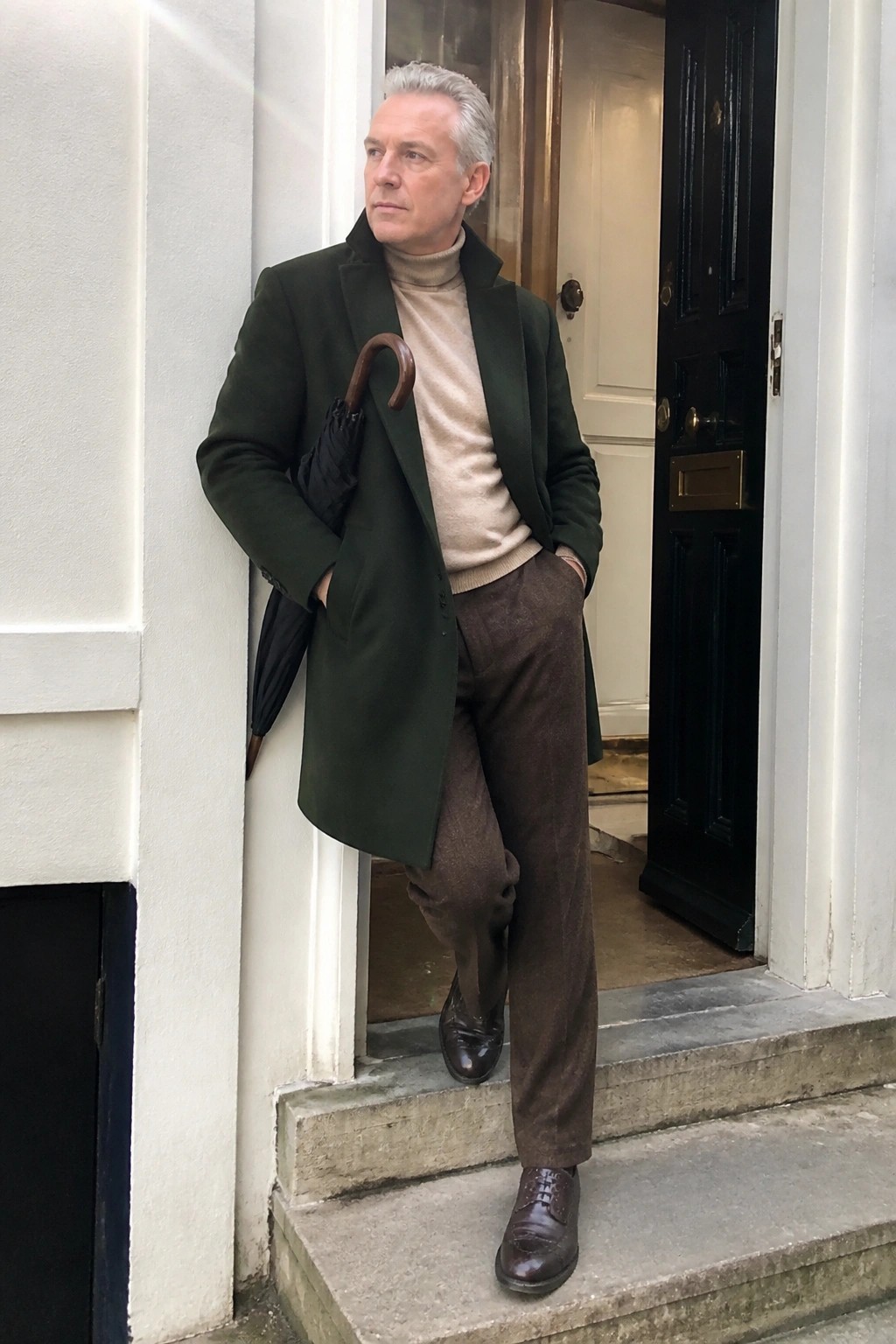 Gray-haired man in green overcoat and beige turtleneck holds umbrella at doorway
