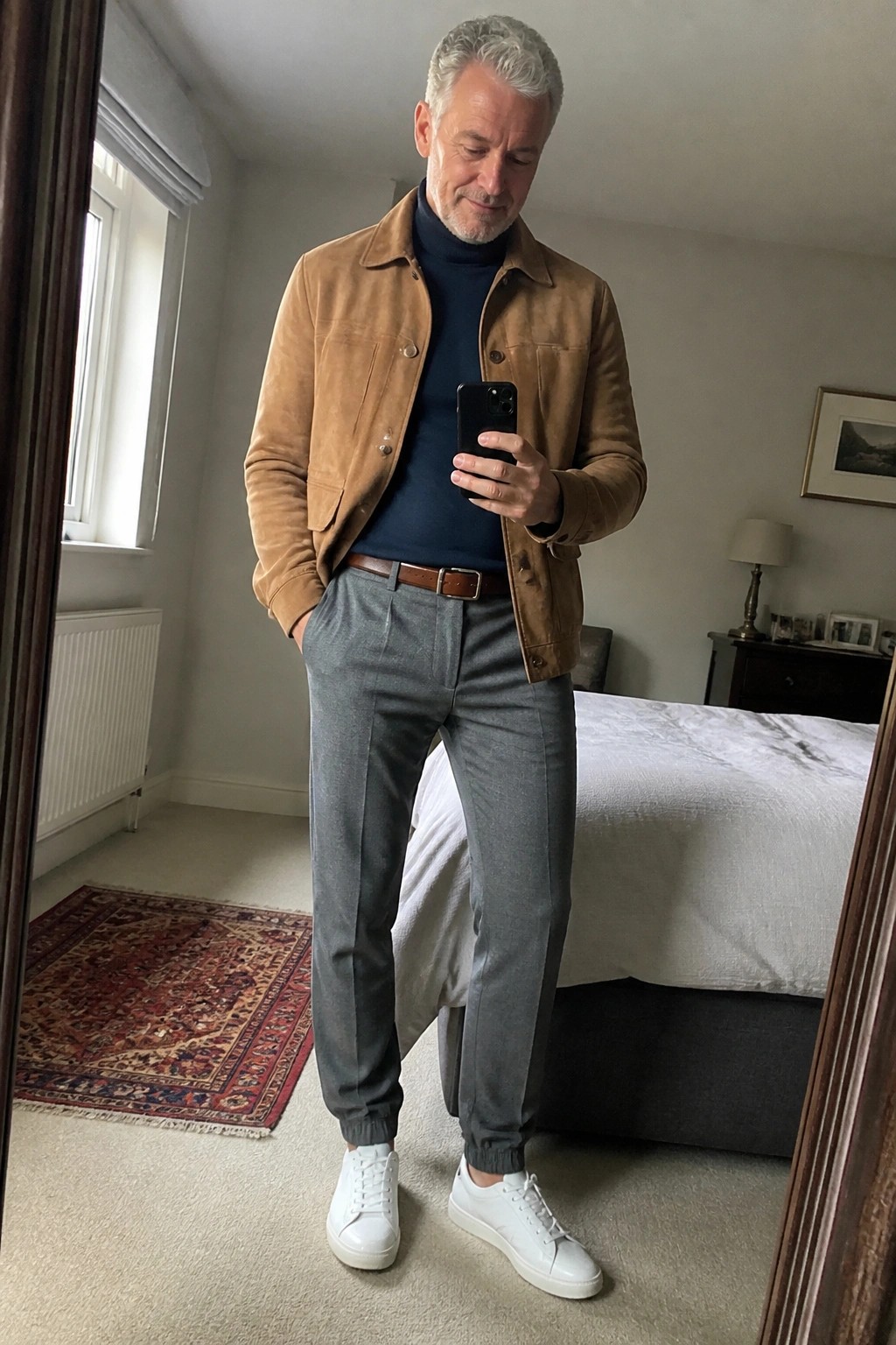Man in brown suede jacket over navy turtleneck checks phone in bedroom