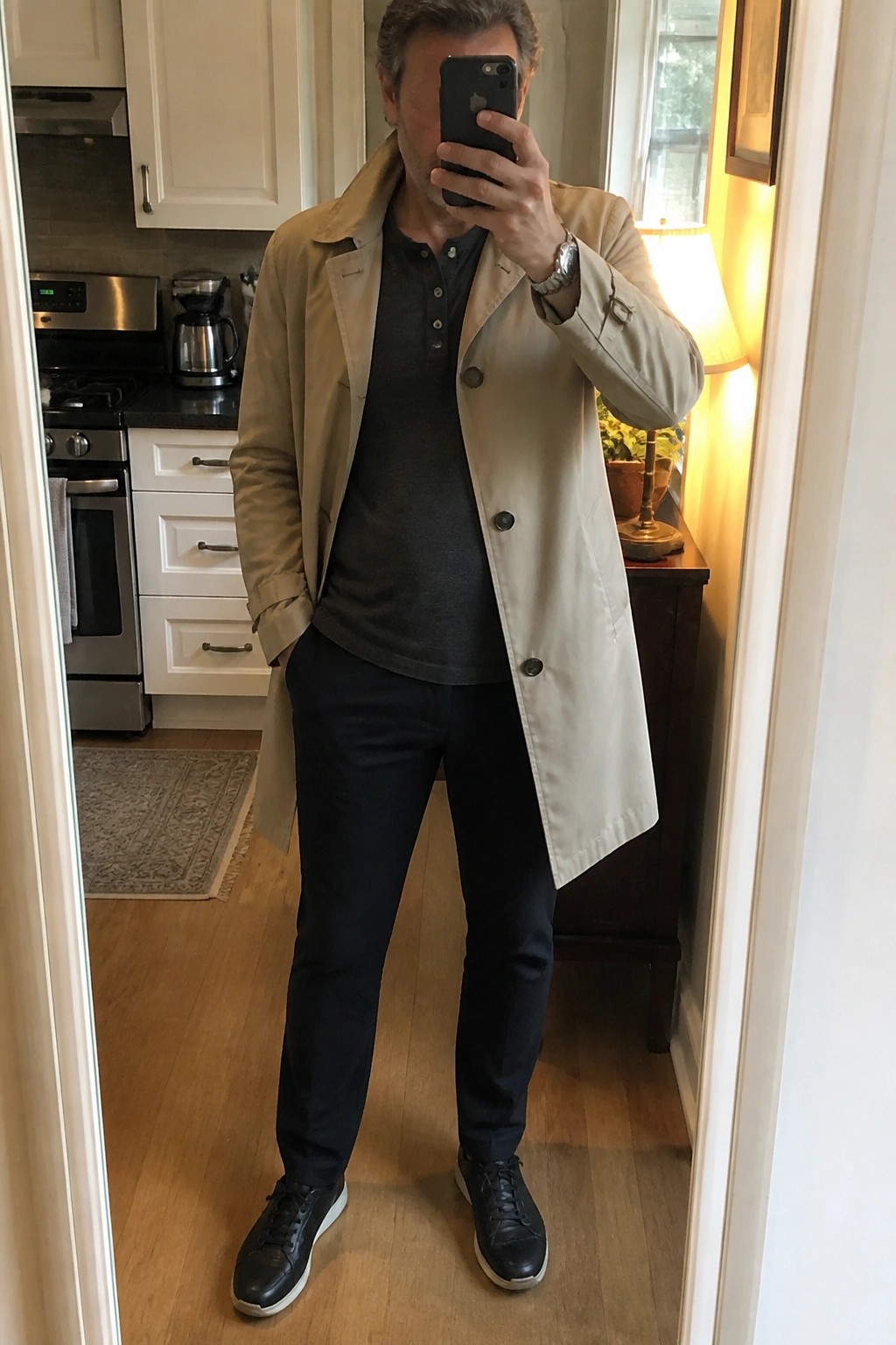Man in a beige trench coat open over a dark gray henley shirt with black pants and black sneakers taking a mirror selfie in a kitchen setting.