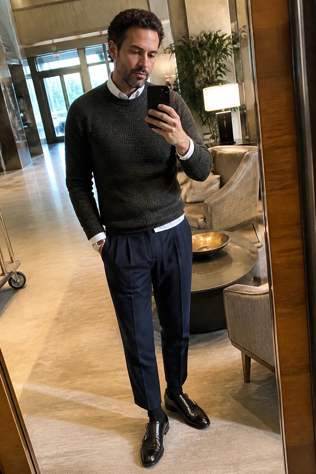 Man in dark knit sweater and navy trousers taking mirror selfie in luxury lobby.
