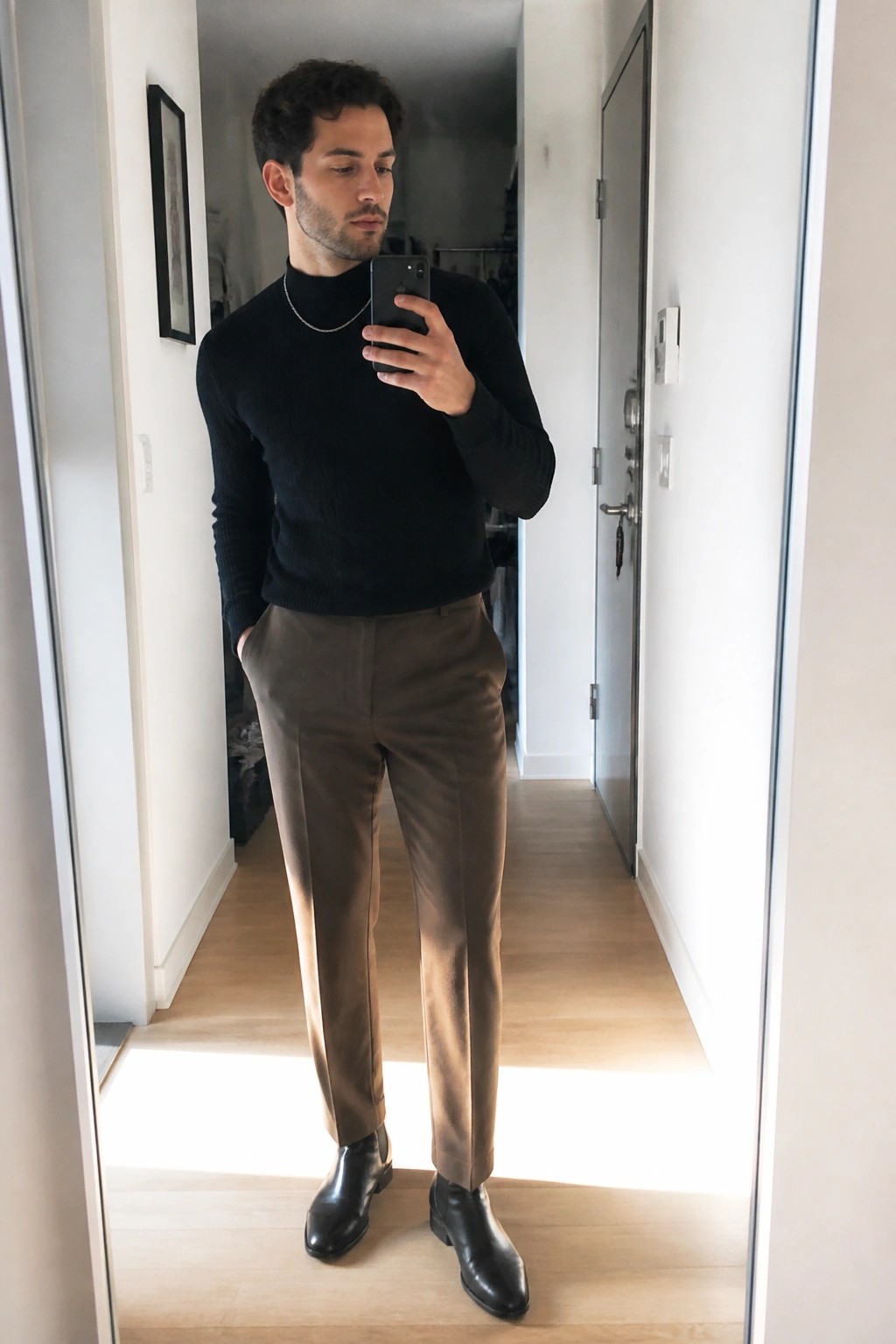 Man in black turtleneck, brown trousers, and black boots taking mirror selfie.