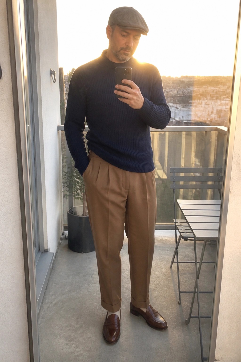 Man in navy sweater, brown trousers, flat cap taking selfie on balcony at sunset
