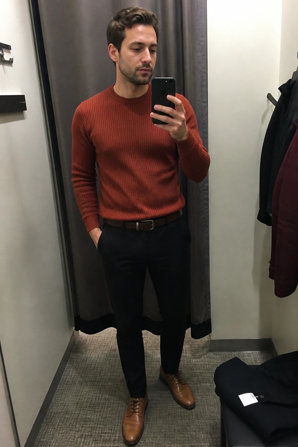 Man in orange ribbed sweater taking mirror selfie in fitting room with black pants
